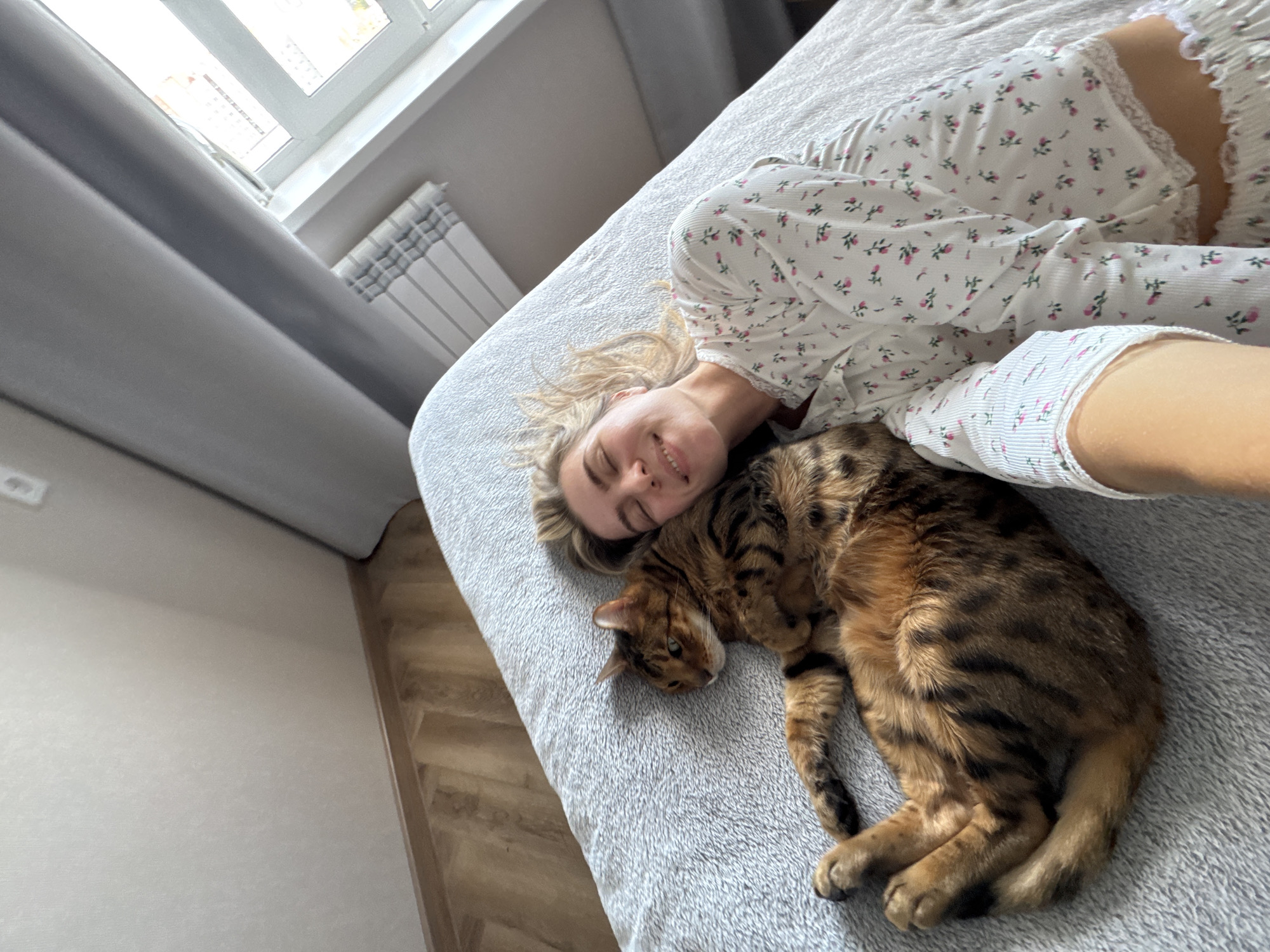 The image depicts a heartwarming indoor scene featuring a woman and a Bengal cat relaxing together on a bed. The woman, with blonde hair, is lying on her side, smiling softly towards the camera, conveying a sense of peace and contentment. She is wearing a light-colored top, possibly sleepwear or loungewear, adorned with a delicate pink floral pattern and lace trim.
Cuddled next to her is a beautiful Bengal cat, distinguished by its vibrant golden-brown fur with distinct dark spots and stripes, typical of the breed. The cat is curled up, appearing to be asleep or resting comfortably on the soft, grey textured blanket that covers the bed.
The setting is a well-lit bedroom, with a large window visible in the background, letting in bright natural light, suggesting it is daytime. Grey curtains are partially drawn, and a white radiator is installed beneath the window. Parts of a light-colored wooden or laminate floor with a subtle herringbone pattern are visible in the corners of the image. The high-angle perspective captures the intimacy and tranquility of the moment. There is no visible text in the image.