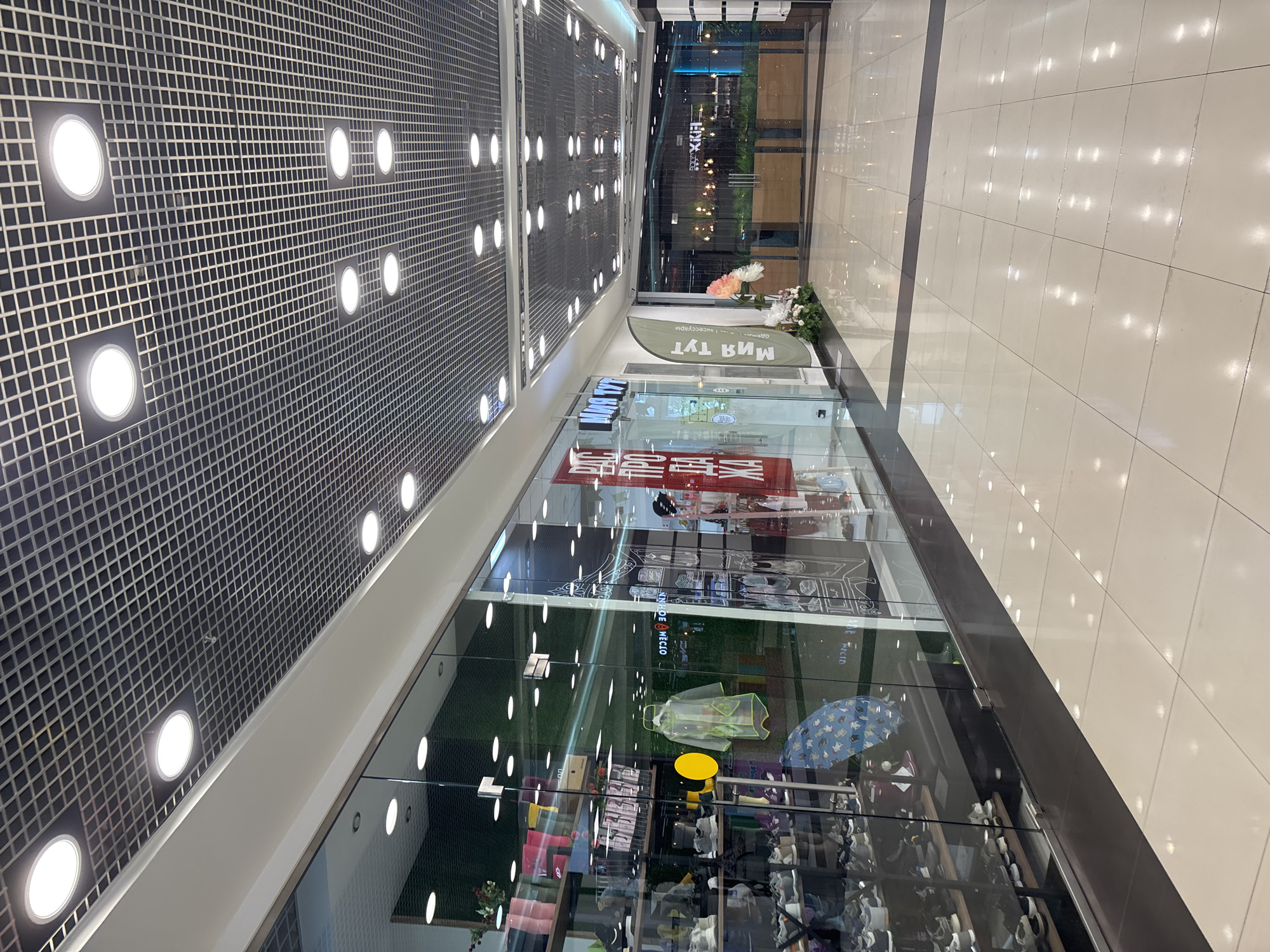 This image shows the interior of a modern shopping mall, specifically a section of a walkway with storefronts and flooring. The ceiling features a grid of recessed LED lights. The floor is tiled with large, light-colored square tiles, reflecting the overhead lighting.
On the left, a storefront with a glass facade displays merchandise on shelves, primarily shoes and accessories. A clear plastic raincoat is hanging on display. To the right of this store is another storefront with Russian signage that includes "МИЯ ТУТ" (Miya Tut - Miya is here). A green flag with similar signage is placed outside this store. Further down the walkway, there are more storefronts, one with a large red sign with Cyrillic text and another that is less distinct. Beyond the immediate storefronts, the mall walkway extends into the distance, with a glimpse of other shops and decorative elements, including a planter with flowers. The overall impression is of a clean, well-lit, and contemporary commercial space.
The scene is identified as being in the city of Naberezhnye Chelny, Russia, suggesting this is a typical commercial environment within that location. There are no people visible in the image, and the focus is on the architecture, interior design, and retail displays of the shopping mall.