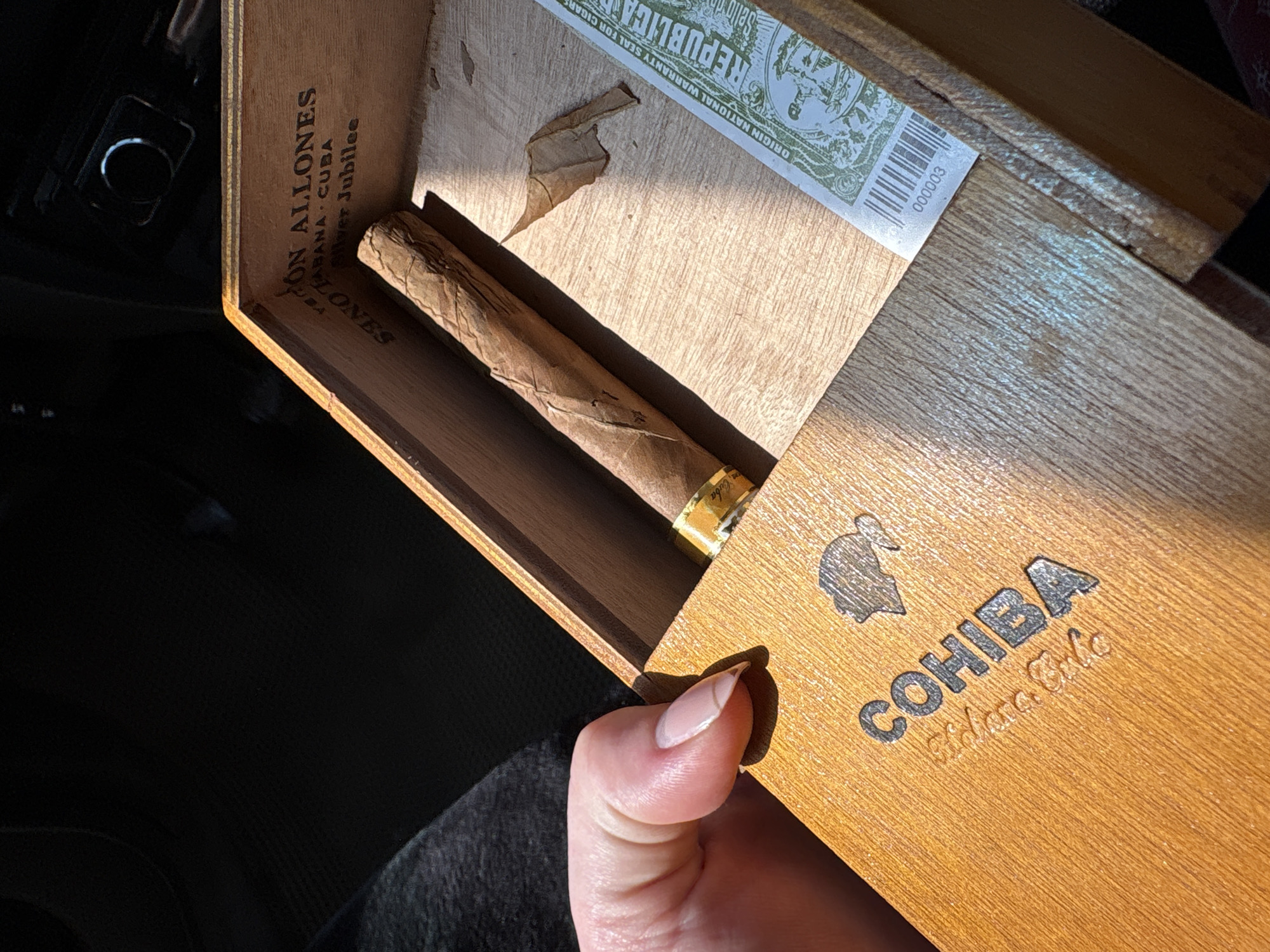 Here is a description of the image:
A partially opened wooden cigar box displays a cigar with a gold band in the center. The interior of the box has an official seal. The box is labeled "ON ALLONES" and "HABANA, CUBA" along the top edge. The box cover is labeled "COHIBA" and is being held open with a hand, a manicured thumbnail is visible. The scene is illuminated by sunlight. The location of the scene is not Фоки, Russia.