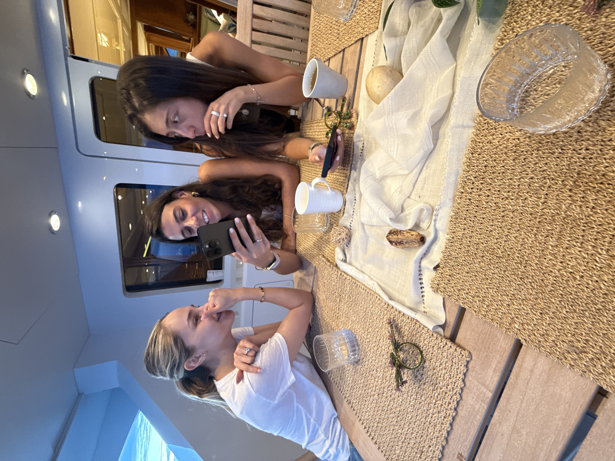 Three young women are seated at a wooden table on a boat, potentially in a marina or on the water, though no specific location within Maravia, Mexico, can be determined from the image. Two of the women are leaning in, looking at a black smartphone being held by one of them, who is smiling. The third woman, closer to the camera, has her hand near her face in a pensive or expressive gesture. The table is covered with woven placemats, a white runner, and various items including mugs, a clear glass bowl, a decorative ball, and a napkin ring. The boat interior is visible in the background, with windows and paneling.