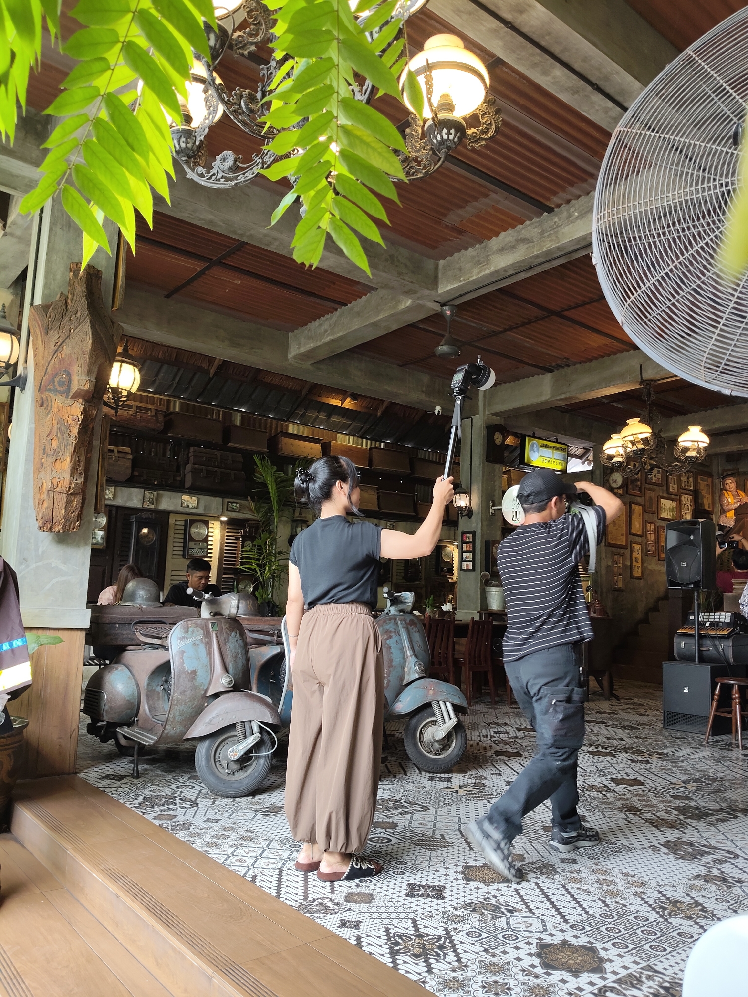 This image depicts the interior of a cafe, possibly named "Jamu Pijat Semarang" as indicated on a screen in the background, located in the City of Medan, Indonesia. The cafe has a rustic and vintage aesthetic, with exposed concrete beams, a corrugated metal ceiling, and antique decorations on the walls, including framed pictures and clocks. Two vintage scooters are prominently displayed on a tiled floor with a patterned design.
In the foreground, a woman wearing a black t-shirt and brown wide-leg pants is holding a selfie stick with a camera attached, seemingly filming or taking pictures. To her right, a man in a black and white striped t-shirt and black pants is adjusting a cap and holding a camera over his shoulder, appearing to be taking photos as well. Several other individuals are visible in the background, some seated and others standing, contributing to the atmosphere of a busy establishment. The lighting in the cafe comes from ornate chandeliers and wall lamps, creating a warm ambiance. A large industrial fan is mounted on the ceiling on the right side of the image. The overall scene suggests a social gathering or a photoshoot taking place within the cafe.