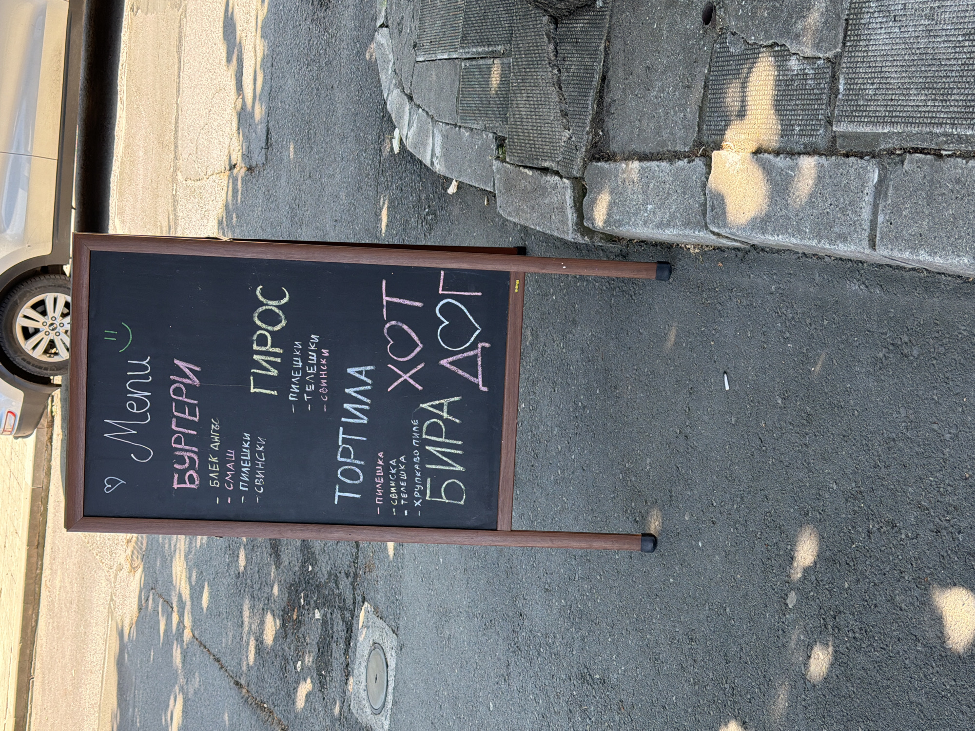 The image shows a black chalkboard menu sign displayed outdoors on a paved surface. The sign is framed with dark wood and stands on two legs. The menu is written in chalk and appears to be in Bulgarian.
The top of the menu reads "Menu" with a heart symbol and a smiley face drawn next to it. Below this, the main categories are:
* "БУРГЕРИ" (Burgers)
* "ГИРОС" (Gyros)
* "ТОРТИЛА" (Tortilla)
Under "БУРГЕРИ", there are bullet points listing:
* "БЛЕК АНГЪС" (Black Angus)
* "СМАШ" (Smash)
* "ПИЛЕШКИ" (Chicken)
* "СВИНСКИ" (Pork)
Under "ГИРОС", there are bullet points listing:
* "ПИЛЕШКИ" (Chicken)
* "ТЕЛЕШКИ" (Veal)
* "СВИНСКИ" (Pork)
Under "ТОРТИЛА", there are bullet points listing:
* "ПИЛЕШКА" (Chicken)
* "СВИНСКА" (Pork)
* "ТЕЛЕШКА" (Veal)
* "ХРУПКАВО ПИЛЕ" (Crispy Chicken)
At the bottom of the menu, "БИРА" (Beer) is written with a heart symbol, and next to it "ХОТ ДОГ" (Hot Dog) is written with another heart symbol.
The menu is placed on asphalt, with a section of concrete curb and paving stones to the right. The background shows a blurred view of a street and a parked car. The overall impression is a casual outdoor dining or takeaway establishment.
The mention of Tsarevo, Bulgaria, suggests this scene is located in that town.