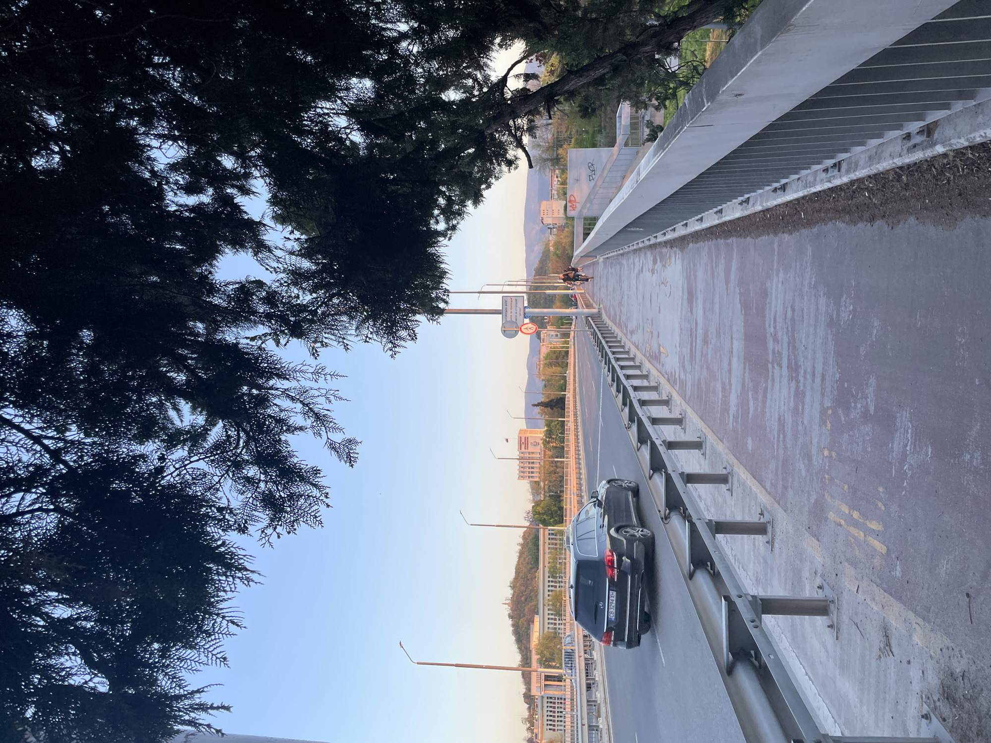 This image, taken in Plovdiv, Bulgaria, captures a multi-lane bridge or overpass during the golden hour, with the sun casting soft, warm light across the scene. The clear sky transitions from a light blue overhead to warmer hues near the horizon.
In the foreground and upper left, dense, dark green coniferous tree foliage partially frames the view, suggesting the photo was taken from beneath or beside a large tree. The bridge extends from left to right, showing a road with at least two lanes for vehicular traffic on the left, and a separate pedestrian or cycling path on the right, divided by a metal crash barrier.
On the vehicle road, a dark-colored car is visible, bearing the Bulgarian license plate "CB 3296 XH". Further along the pedestrian/cycling path, a person on a motorcycle or scooter is seen. Several streetlights are positioned along the bridge structure. A white road sign, partially obscured, displays Cyrillic text including "ВНИМАНИЕ НАЙ" (ATTENTION MOST) and possibly "И В ГРАДА" (AND IN THE CITY), alongside a circular red traffic symbol (likely a 'no entry' variant).
A concrete wall running along the right side of the bridge features graffiti, with the words "FLER" and "WDP" clearly visible. In the mid-ground and background, the urban landscape of Plovdiv unfolds, with various buildings nestled among gently rolling, vegetated hills. Distant mountains complete the horizon under the soft light of late afternoon or early evening. The scene conveys a tranquil moment in an urban environment at dusk.