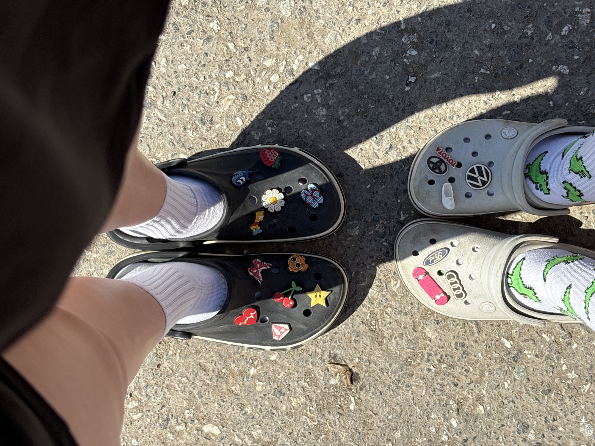 The image depicts two pairs of Crocs shoes on a concrete surface, possibly a sidewalk or pavement. One pair is black and adorned with decorative charms, including a strawberry, a daisy, and a butterfly. The person wearing these Crocs is wearing white socks. The other pair is light grey, also embellished with charms of automobile brands such as Volkswagen, Toyota, Ford, and Audi, and a skateboard. This person is wearing white socks with green lizard images. The scene is outdoors, evidenced by the sunlight and shadow cast across the concrete. There is no indication of the location being in Grishanki, Russia, as no specific geographical or cultural markers are present.