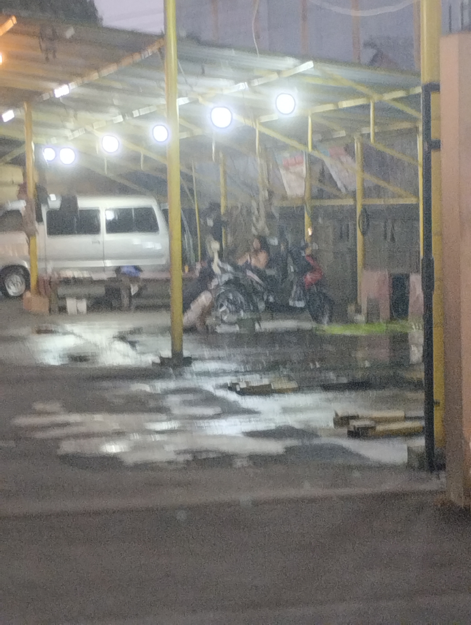 The image depicts an outdoor parking area, likely a car wash or service station, under a yellow metal awning supported by yellow poles. The ground is wet and reflects the overhead lights, suggesting recent rain. Several vehicles are present, including a white van parked on the left, and multiple motorcycles parked in the center and right. Some people are visible, appearing to be working on or near the motorcycles. In the background, there are structures with windows and doorways, indicative of a commercial area. The scene appears to be set in the city of Bekasi, Indonesia.