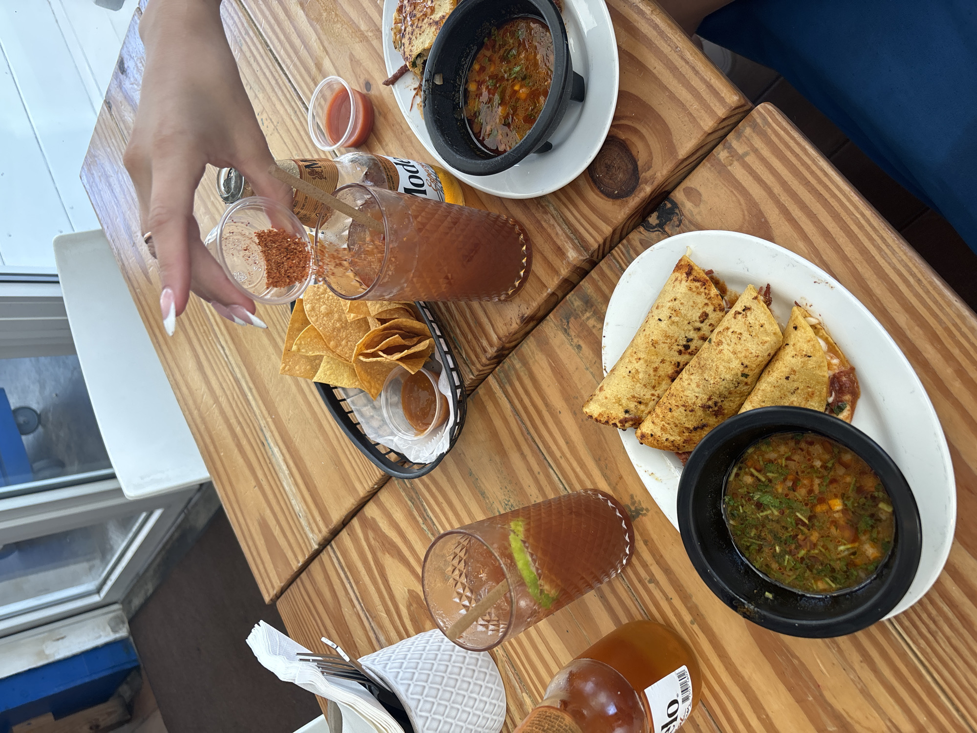 The image shows a close-up, overhead view of a table laden with Mexican food and drinks. A person's hand with manicured nails is reaching to add chili powder from a small container into a glass of reddish liquid. The food includes three golden-brown quesadillas on a white oval plate, a black bowl filled with a chunky salsa, and a basket of tortilla chips with a small cup of salsa. There are two glasses of drinks on the table; one is a reddish beverage with lime and a straw, and the other appears to be the same drink into which chili powder is being added. A bottle of Modelo beer is also visible, along with a small bottle of hot sauce. The setting is a casual outdoor or indoor dining area with a wooden table and a glimpse of a white and blue structure that could be a window frame or part of a building. The natural light suggests it is daytime, likely afternoon, with no specific weather cues. The overall impression is of a relaxed meal, possibly lunch or an early dinner, with friends or family. The focus is on the food and beverages, highlighting the ingredients and preparation. The visible text on the beer bottle is "MODELO" and on the hot sauce bottle, part of a label is visible.