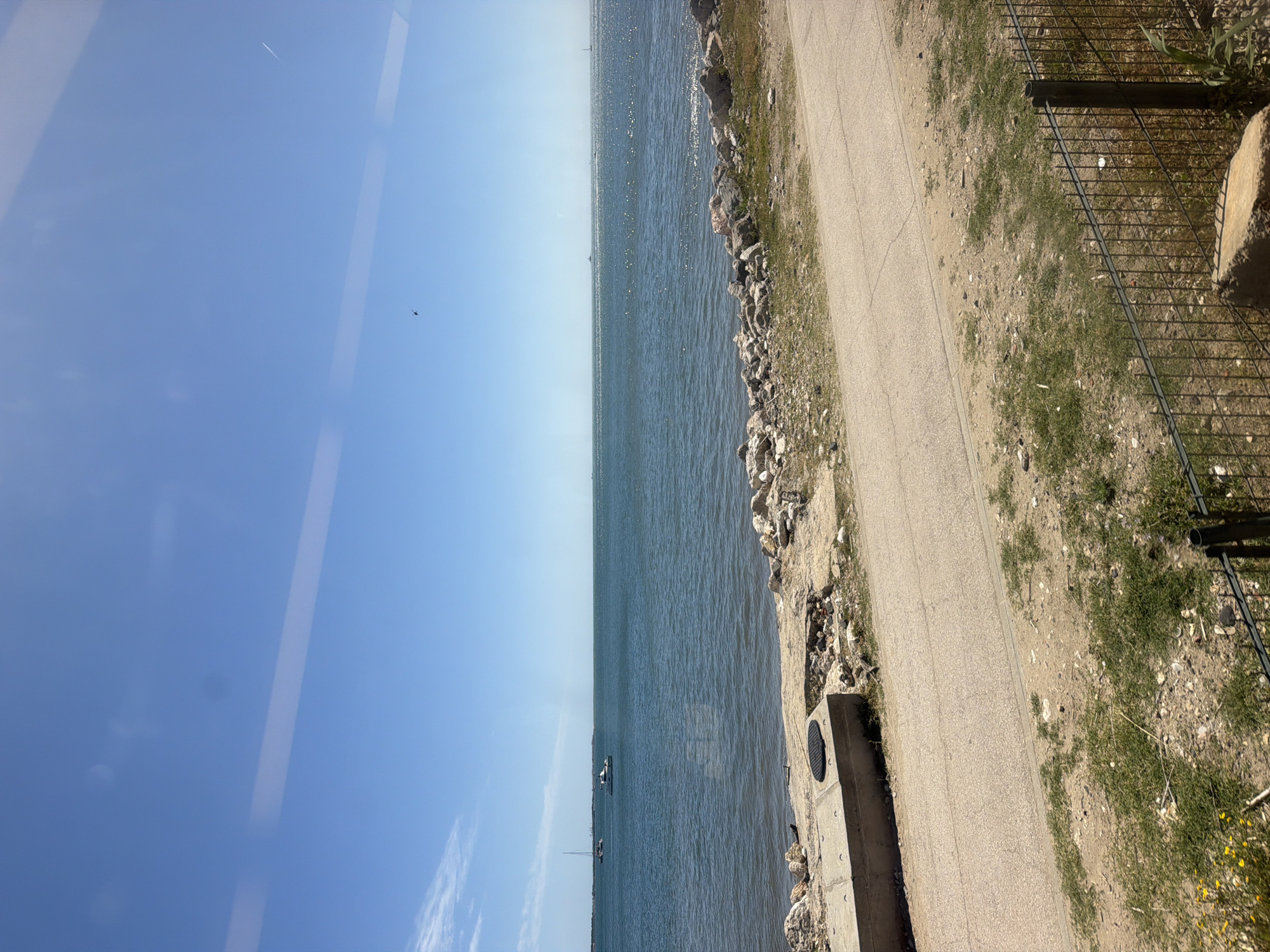 A coastal view in Oeiras, Portugal. The scene captures a narrow paved pathway running alongside a rocky shoreline and the Tagus estuary. A metal fence separates the pathway from a grassy area. The water is calm with a few boats visible in the distance. The sky is clear blue. The image appears to be taken through a window.