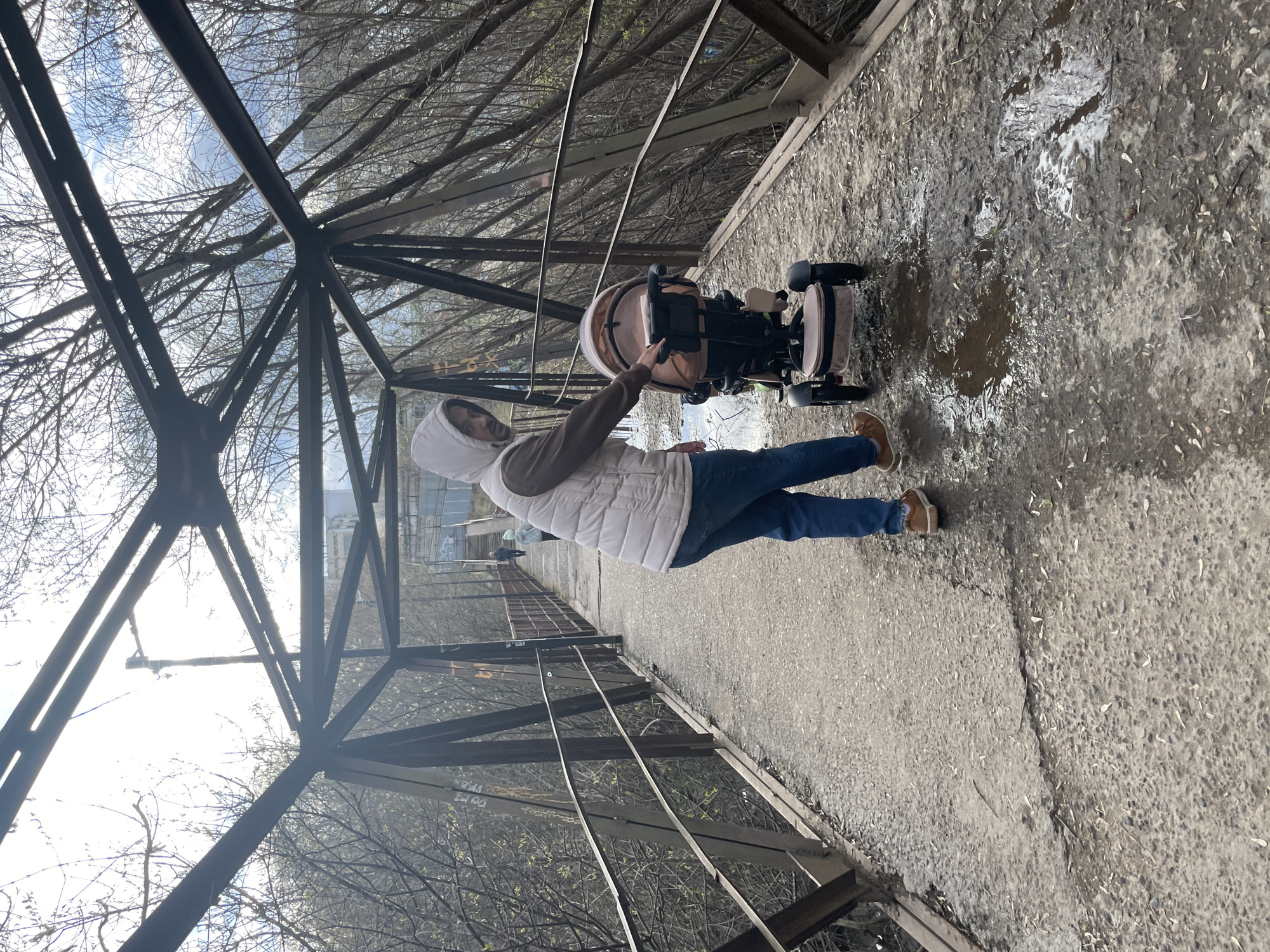 The image captures an outdoor scene on what appears to be an elevated walkway or bridge in Izhevsk, Russia. The sky above is cloudy with bright patches of daylight, visible through a dense canopy of bare tree branches and a lattice of dark metal structural beams.
In the foreground, an adult person stands on a wet, paved pathway. They are dressed in a light-colored hooded vest over a long-sleeved shirt, blue jeans, and brown shoes. The person is positioned next to a beige and black child's stroller or tricycle, with one hand extended towards it, suggesting they are about to push or adjust it. The pathway itself is uneven, with several puddles and scattered debris, indicative of recent rain or melting conditions.
The walkway is framed by a heavy metal structure, forming both an overhead support and side barriers. Beyond these structures and trees, faint outlines of distant buildings can be discerned. On one of the metal beams forming the side barrier, there is white graffiti, with some legible Russian characters appearing to be "РОЙ ЕЛ" or "РОЙ ЕЗ".