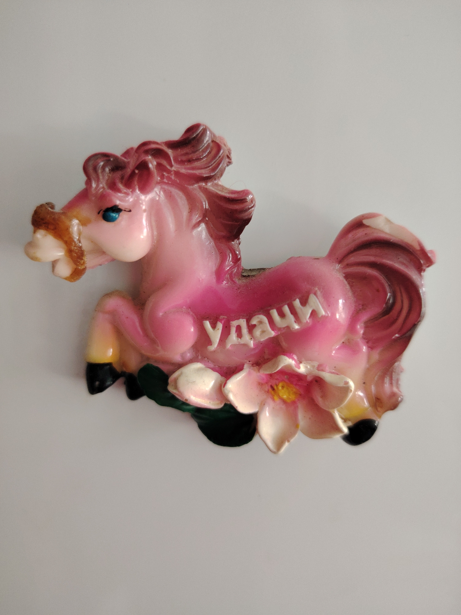 A pink, glossy decorative figurine of a horse is depicted against a plain light background. The horse is in a dynamic, rearing pose, with its front legs lifted. It features blue eyes and a darker pink/magenta mane and tail. At the base of the figurine, there are two light-colored flowers with yellow centers and some green leaves. The word "УДАЧИ" (UDACI), which translates to "Good Luck" in Russian, is inscribed in raised white capital letters on the side of the horse's body. The object appears to be a magnet or small ornament, possibly made of ceramic or resin, and shows some minor signs of wear and dust. The location associated with this media is Сусады-Эбалак, Russia.