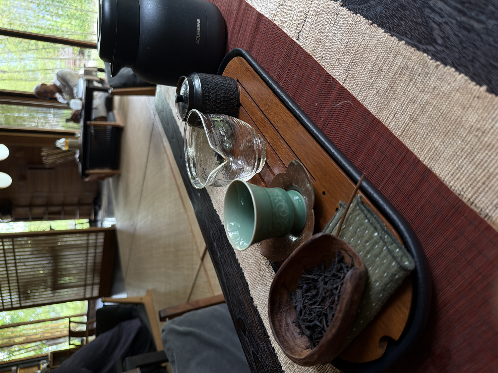 A still life photograph taken indoors in Sukawati, Indonesia, depicts a tea set arranged on a wooden tray with a black rim. The tray rests on a striped table runner featuring beige, maroon, and black stripes. The tea set includes a black electric kettle, a textured black filter, a clear glass pitcher with a spout, a light green teacup resting on a saucer, a wooden bowl containing dark tea leaves, and a small square green cloth with white dots. In the background, a blurred view through a window reveals a person sitting at a table, other indistinct objects, and a lush green outdoor environment. The composition suggests a moment of tea preparation or enjoyment.