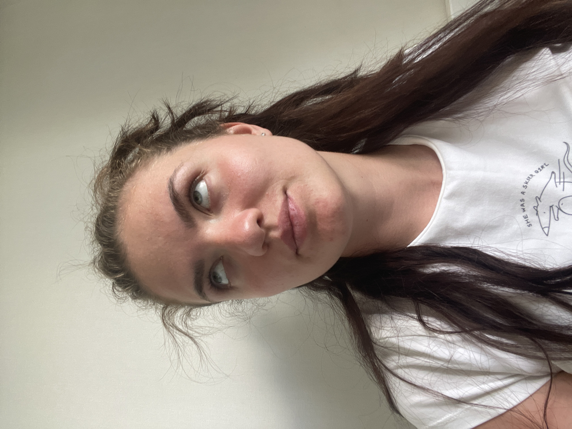 A young woman with fair skin, dark brown hair, and light-colored eyes is visible in the image. She is wearing a white t-shirt with a printed design, part of which reads "SHE WAS A SKIER GIRL". The background appears to be a plain, light-colored wall. The photo seems to be taken indoors. The location is not discernible within Moscow, Russia, or anywhere else, based on the provided image.