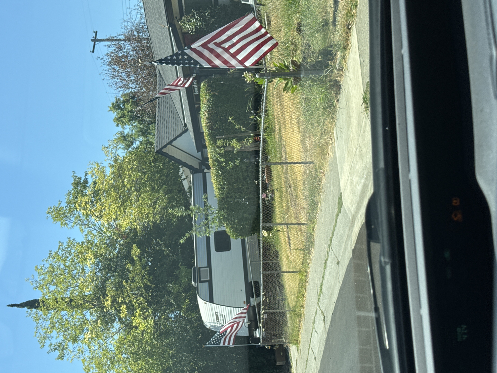 The image shows a white recreational vehicle parked in a residential yard in Citrus Heights, California. Two American flags are planted in the yard, one near the front of the RV and another in front of a house in the background. The house appears to be a single-story dwelling with a dark roof. A chain-link fence separates the yard from the street. Trees and bushes are visible in the background, and the sky is clear and blue. The photo appears to be taken from inside a vehicle, with the dashboard and windshield visible in the foreground.
