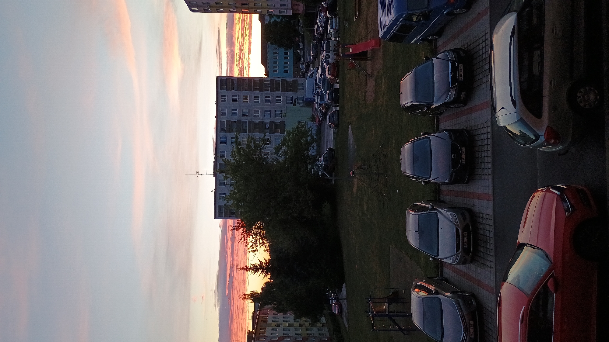 The image captures a sunset view from a high vantage point, likely an apartment building, in Sokolov, Czechia. The sky is a canvas of soft pinks and oranges, transitioning into a pale blue, indicating the time of day is dusk. In the foreground, several cars are parked on a paved area adjacent to a grassy lawn. These include a red car, a silver car, and two dark-colored cars. Beyond the parking area, a multi-story residential building with numerous windows is visible. Further in the background, other buildings of varying heights and colors, including a pale blue one, are present. Trees and a children's playground with a red slide are also visible on the lawn. The overall scene suggests a quiet evening in a residential neighborhood. No people are visible in the frame. Some license plates are partially visible on the cars, but the text is not legible.