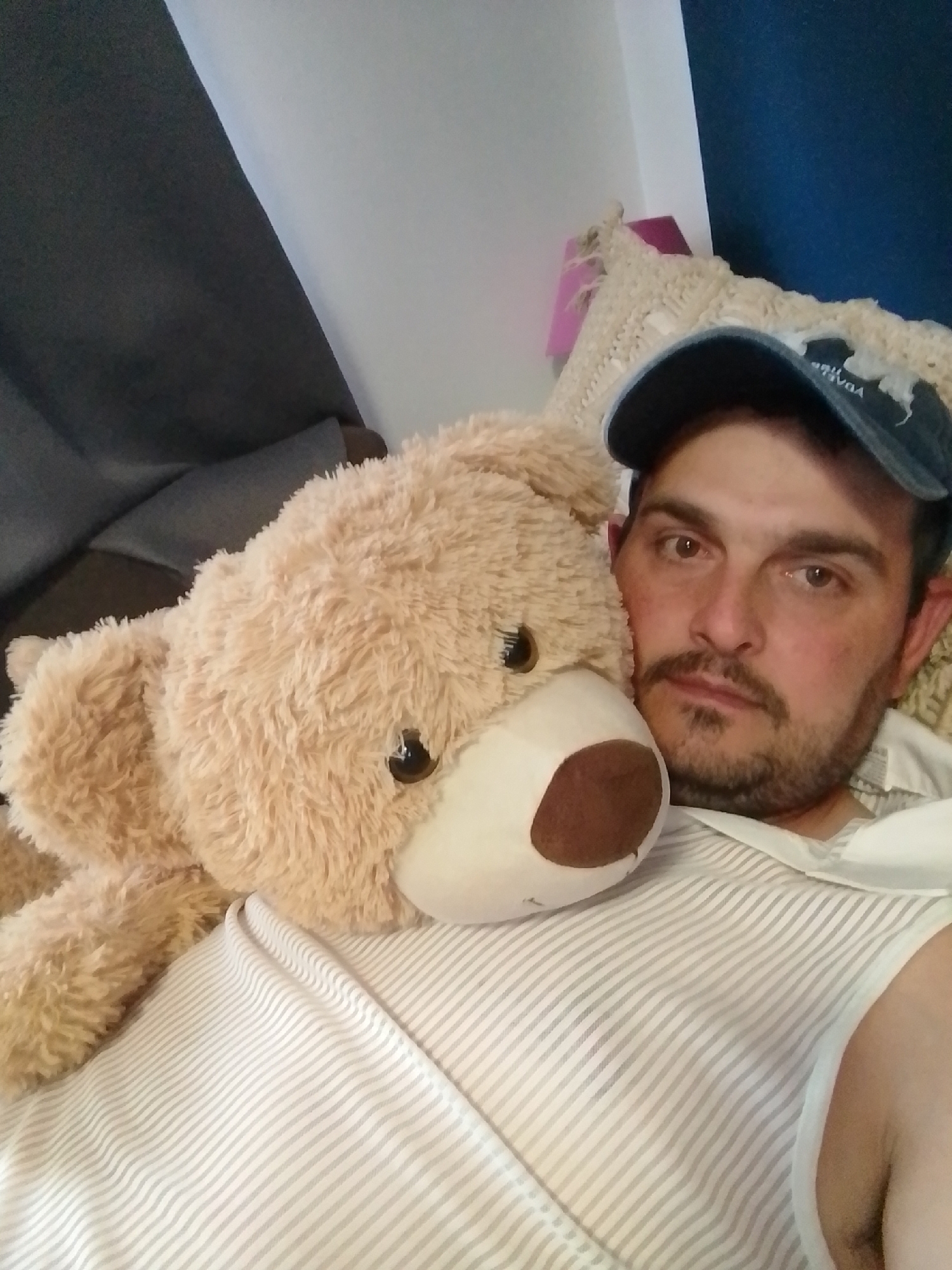 In Wedel, Germany, an individual lies on a bed, embracing a large teddy bear. The person, wearing a blue baseball cap and a striped shirt, has their face partially obscured by the bear. The room appears to be a bedroom, with a dark gray couch visible in the background and a blue wall behind. The overall atmosphere is relaxed and personal, suggesting a moment of quiet repose.