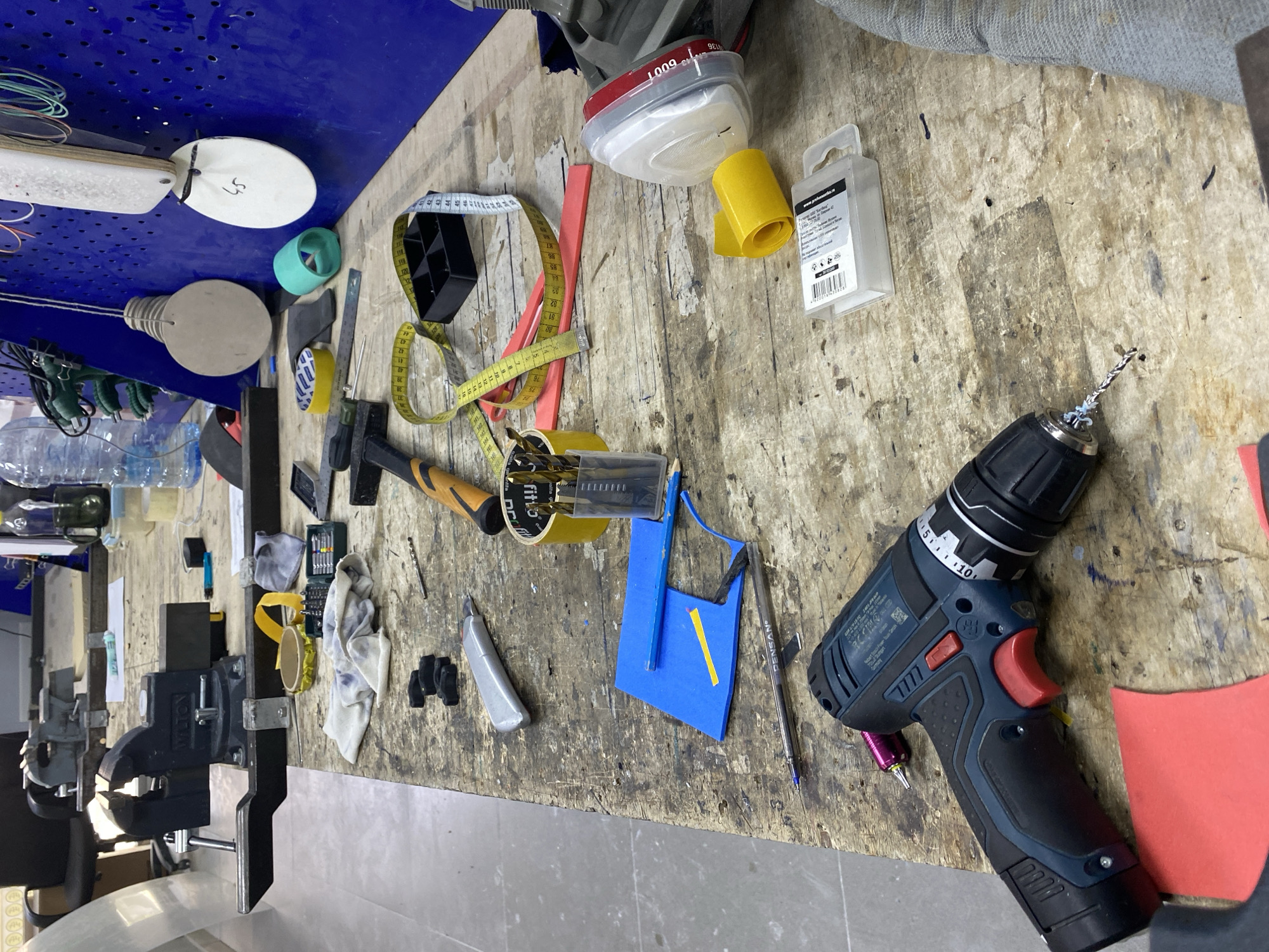 This is a close-up, eye-level shot of a cluttered wooden workbench in what appears to be a workshop or garage, possibly located in Vladimir, Russia. The workbench is covered with a variety of tools and materials, suggesting active work is in progress or has recently concluded.
Prominently featured in the foreground is a blue and black cordless drill with a red trigger and chuck. To the left of the drill, a piece of red material is visible. Further back on the workbench, a silver utility knife is present, alongside several small black objects that resemble miniature car tires or possibly seals. A roll of yellow tape and a yellow dispenser are also visible.
In the mid-ground, a hammer with an orange and black handle rests on the bench. Scattered around it are several drill bits in a clear plastic container, a yellow tape measure, and a black plastic holder with compartments. A blue piece of foam or plastic, cut into a trapezoidal shape with a yellow strip, lies near the hammer. A pen with the brand name "PENSAN" is also on the bench.
In the background, a blue pegboard is mounted on the wall, with various items hanging from it, including circular wooden discs, one with the number "45" written on it, and a teal-colored object. A bottle of water and other items are partially visible in the upper left. The overall impression is one of a functional, albeit messy, workspace. There are no people or discernible actions in the image.
