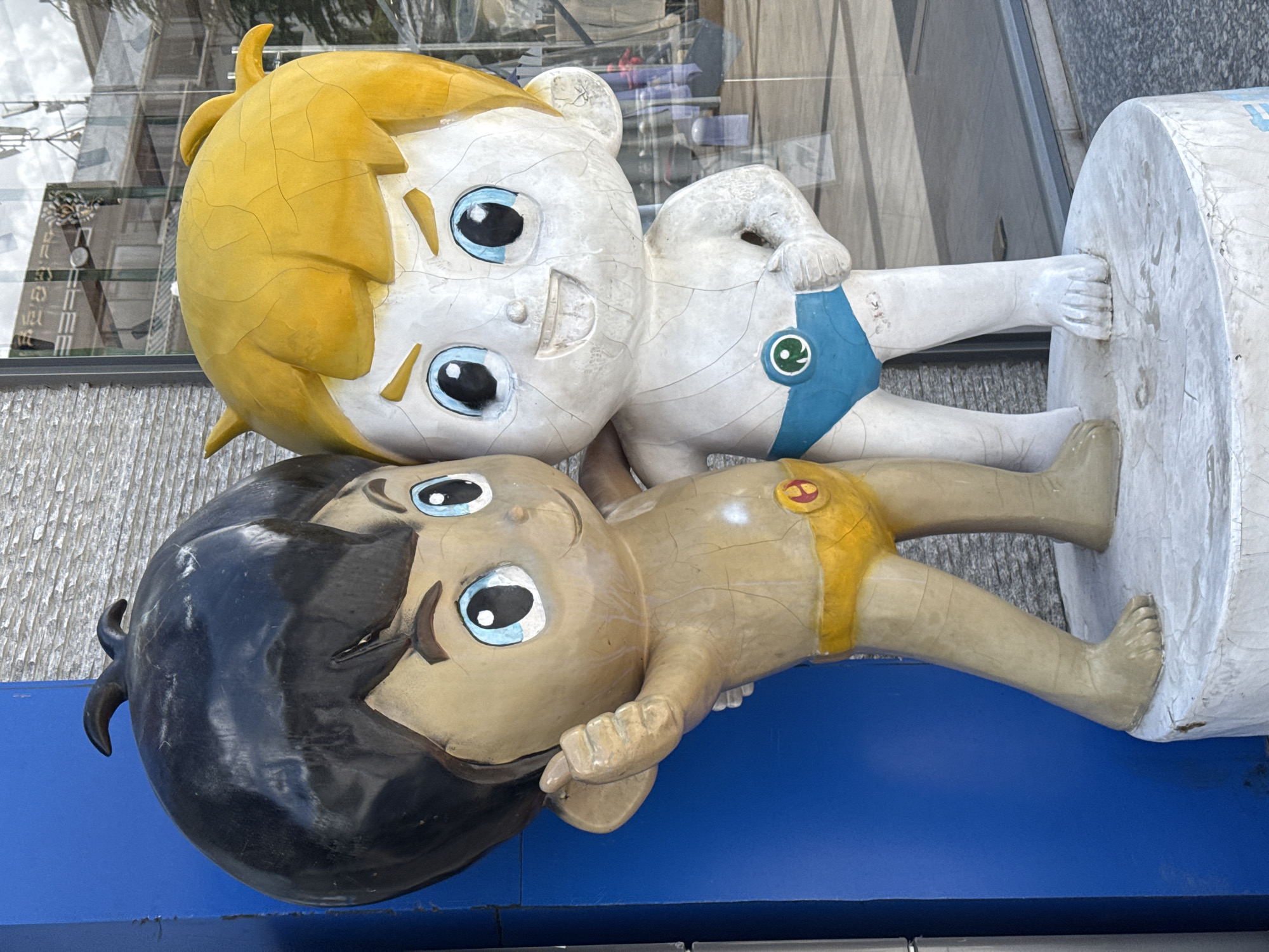 Two cartoon-like statues of children are depicted, appearing to lie side-by-side. The image is oriented vertically, so the figures are arranged one above the other in the frame. The upper statue has bright yellow hair and light-colored skin, with large blue eyes and a cheerful smile. It wears blue shorts featuring a green circular emblem. Its body shows noticeable cracks and wear.
The lower statue has dark hair and tan skin, with similar large blue eyes and a gentle smile. It wears yellow shorts adorned with a red circular emblem. This statue also exhibits significant surface cracks and deterioration. Both figures have their arms bent, with the upper figure's right hand (as viewed from the top of the statue) resting against a white, cylindrical base or pillar that supports both figures. The lower figure's feet also rest against this cylindrical base.
The background shows reflections of modern architectural elements, primarily glass windows of buildings, indicating an urban outdoor setting, possibly a commercial area. The lighting suggests it is daytime. The statues appear to be part of a public art installation or display, possibly mascots, and their weathered appearance indicates exposure to the elements over time. No legible text is visible on the statues themselves or their immediate bases. The overall scene conveys a sense of whimsical public decoration.
