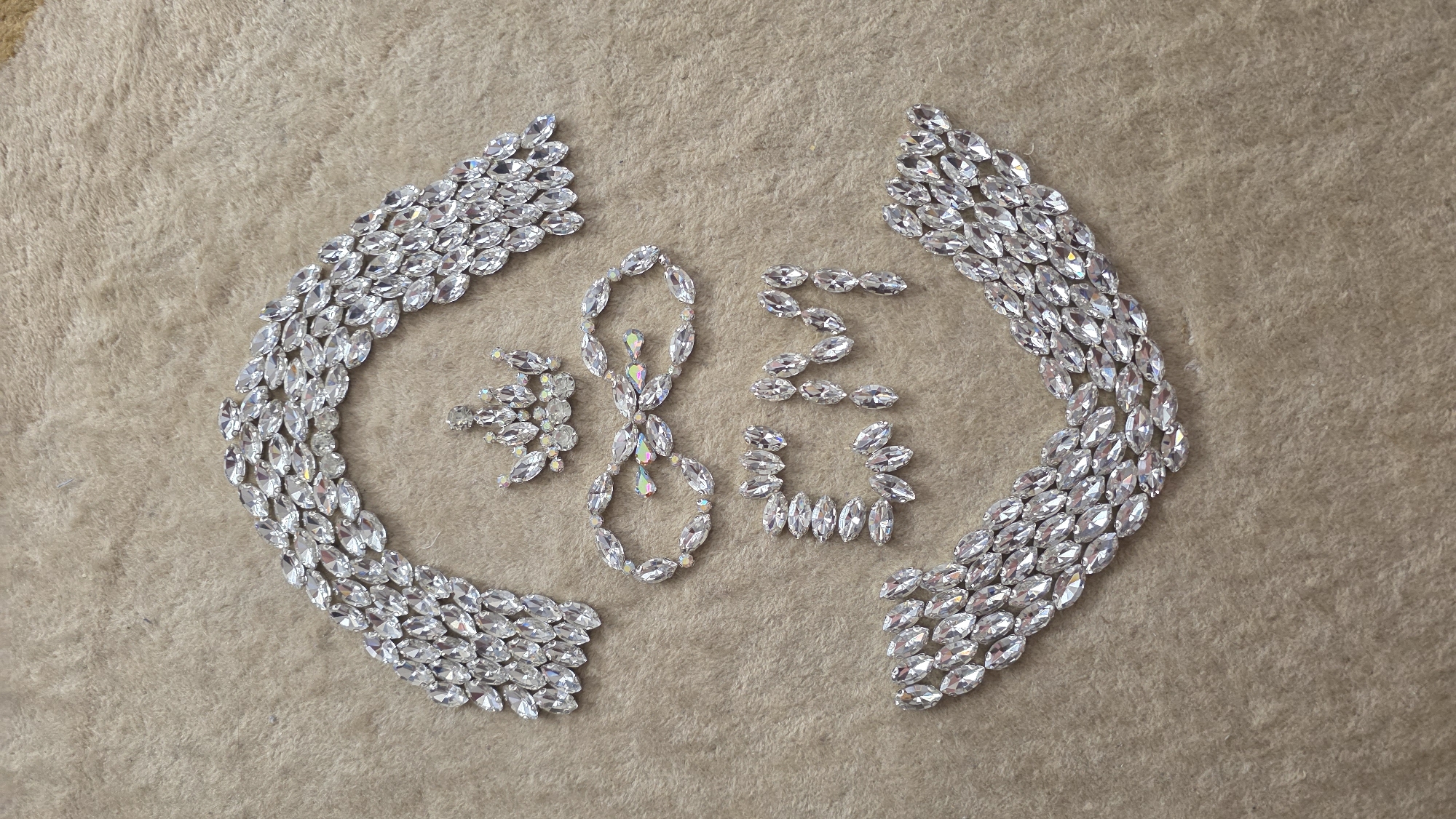 The image captures an artistic arrangement of numerous clear, faceted rhinestones on a textured, light brown surface, likely felt or carpet, in Blagoevgrad, Bulgaria. The sparkling objects are primarily marquise-shaped, with some smaller, round, and iridescent stones interspersed within the central design.
The arrangement is composed of two large, symmetrical, wing-like or bracket-like structures flanking a central display. Each of these outer elements is intricately formed from multiple rows of the clear marquise rhinestones. In the space between these two larger formations, the numbers "2023" are meticulously spelled out using similar rhinestones. The individual digits are distinct, with the "0" and "3" incorporating a few iridescent, multi-colored pieces that reflect light in a prism-like manner, adding a subtle splash of color.
The background surface provides a soft, uniform, and slightly fuzzy texture, accentuating the sharp facets and brilliant sparkle of the rhinestones. The lighting is bright and even, highlighting the reflective qualities of the stones and the precise craftsmanship of the arrangement. No human activity is depicted; the image focuses solely on this decorative, static display, possibly a crafting project or a celebratory emblem for the year 2023.
