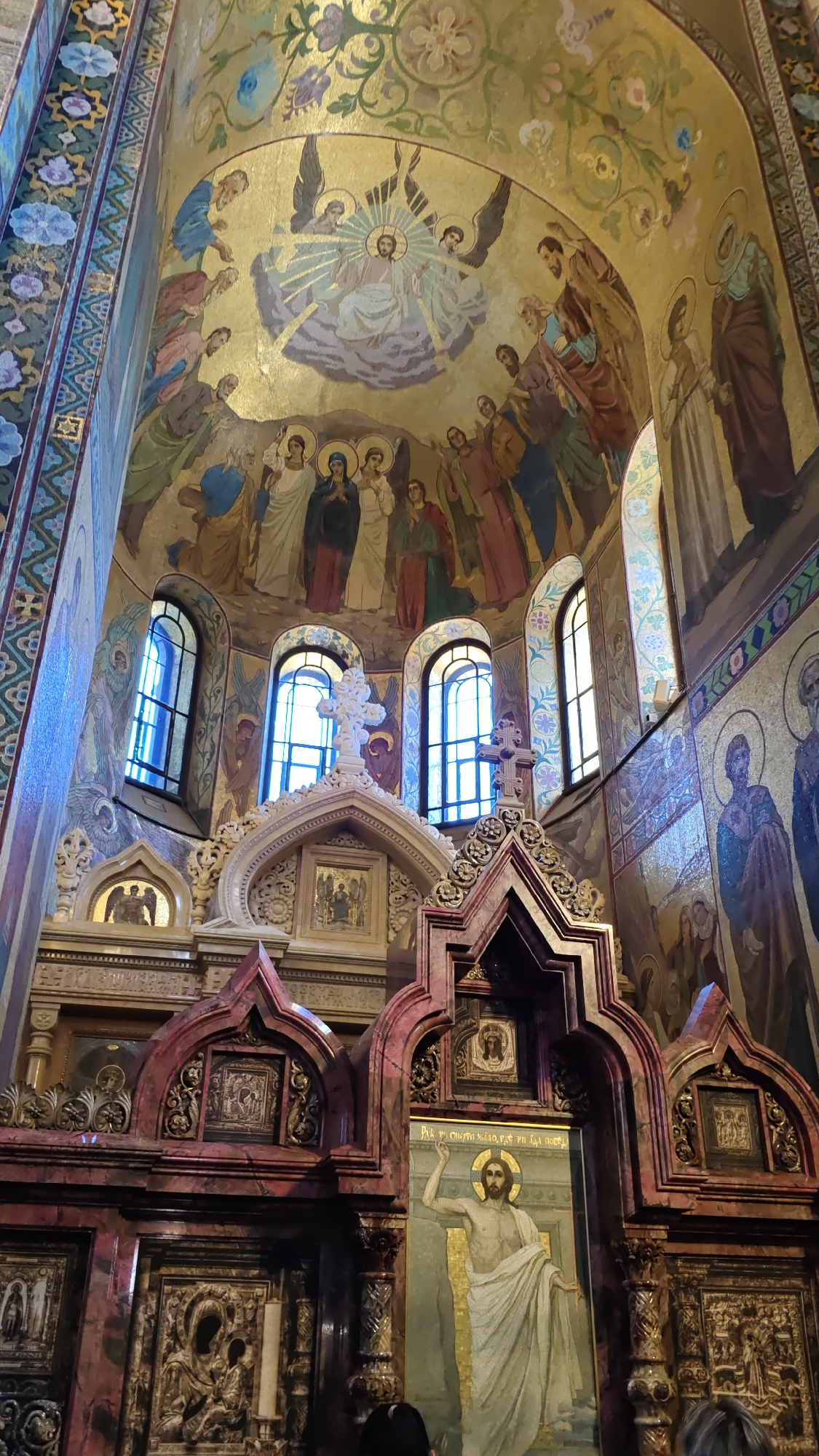 The image captures the interior of the Church of the Savior on Spilled Blood in Saint Petersburg, Russia. The ornate church is adorned with numerous mosaics and frescoes. In the upper portion of the image, a large mosaic depicts Jesus Christ surrounded by angels and a gathering of people, rendered in vibrant colors against a gold background. Below this, arched windows are visible, flanked by further mosaic artwork. The central area features a detailed architectural structure, possibly part of an iconostasis, with decorative carvings and religious imagery, including a large mosaic of Christ on the right side. The overall impression is one of rich religious artistry and grand architectural design.
