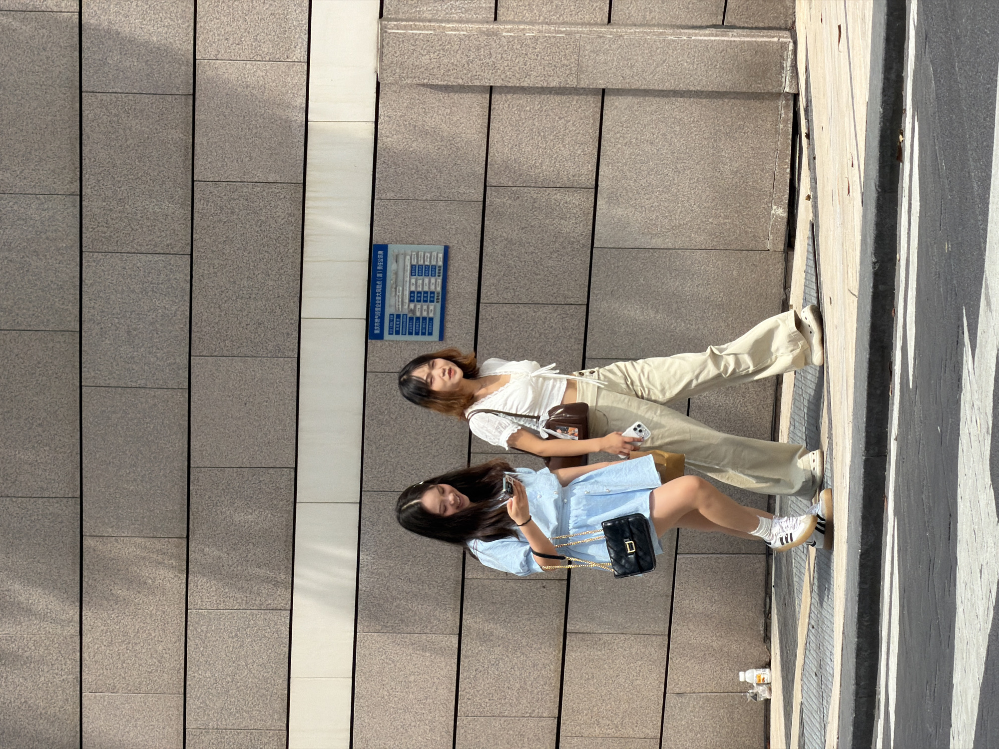 Two young women are walking on a sidewalk in front of a textured stone building. The woman on the left is wearing a light blue dress and carrying a black purse. She is looking at her phone and smiling. The woman on the right is wearing a white crop top and beige pants, carrying a brown purse and a phone. She is looking forward. A blue sign with Chinese text is mounted on the building wall between the two women. The sidewalk is paved with grey tiles and there are some fallen leaves. In the foreground, a pedestrian crossing with white stripes is visible on the asphalt street. The lighting suggests it is a sunny day. The building wall is made of rectangular stone blocks.