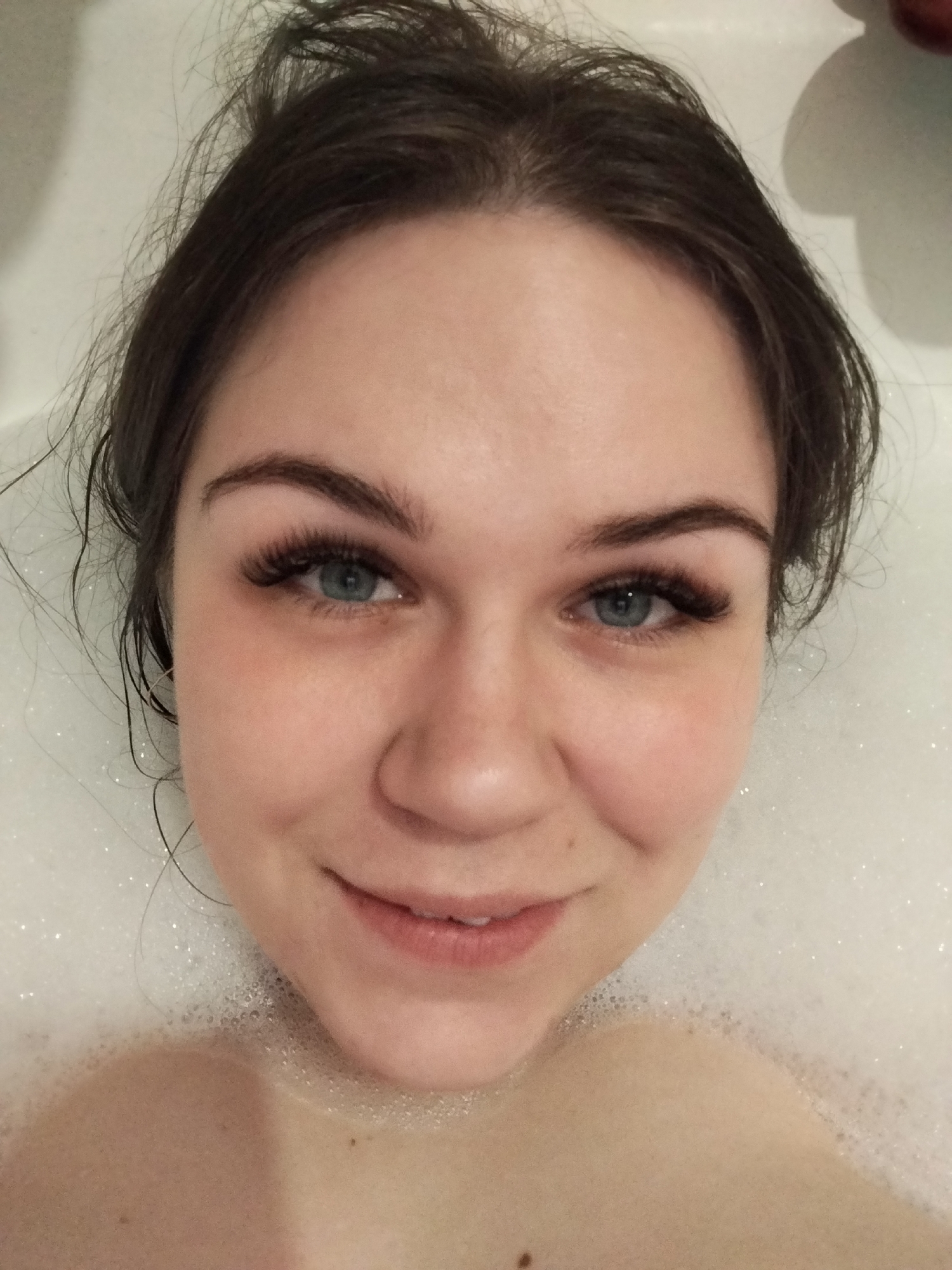 The image shows a close-up, top-down view of an adult female submerged in a bathtub filled with bath foam. The subject has brown hair, blue eyes with prominent false eyelashes, and fair skin. Her facial expression is a gentle smile, and she is looking directly at the camera. Wet strands of hair are visible around her face. Her bare shoulders and upper chest are visible above the white bath foam. The background is the white interior surface of the bathtub. No details within the image allow for the identification of a specific location in the city of Chaikovsky, Russia.