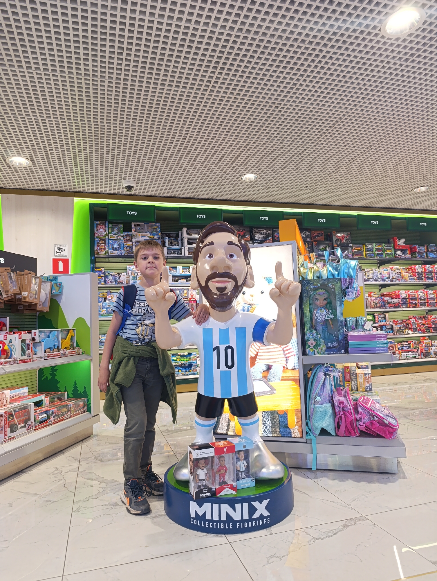 A young boy stands next to a life-sized statue of Lionel Messi in a toy store. The boy is wearing a striped t-shirt, dark pants, and sneakers. He has a green jacket tied around his waist and a backpack on his shoulders. The statue of Messi is wearing his Argentinian national team jersey with the number 10. The store is filled with shelves of toys, with "TOYS" signs visible on the upper shelves. The flooring is tiled with a marble pattern. The image appears to have been taken indoors, possibly in a mall or department store. The specific location in Domodedovo, Russia, is not ascertainable from the image alone but the context suggests a retail environment.