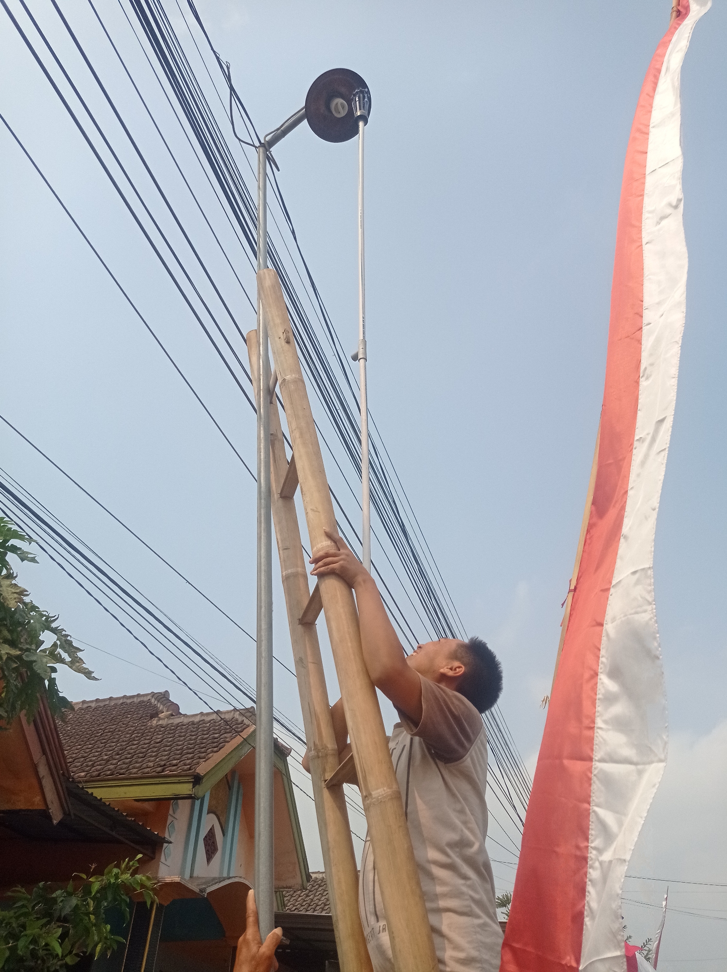 In Purwosekar, Indonesia, a young male with short dark hair is using a bamboo ladder to reach a street lamp attached to a metal pole. He is wearing a grey t-shirt and is gripping the bamboo ladder with his right hand, looking upwards at the lamp. Several dark electrical wires are visible crisscrossing the sky behind him. To the right of the scene, a large red and white Indonesian flag is partially visible, hanging vertically. In the background, parts of houses with tiled roofs can be seen. The sky is a pale blue with a few faint clouds.