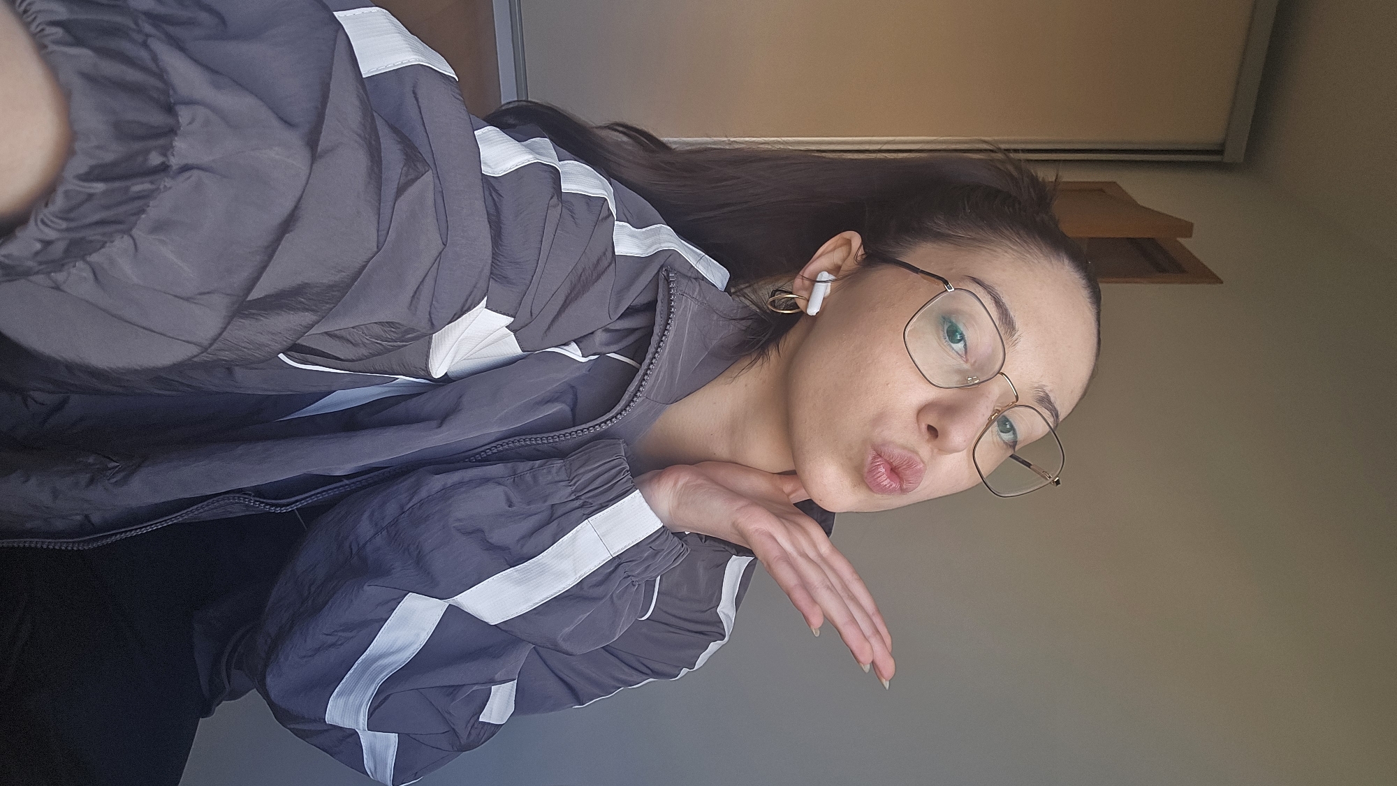 A young woman is oriented at an angle to the right. She is wearing glasses and has earbuds in her ears. She is wearing a gray jacket with white stripes. Her hand is held up near her face, and she is puckering her lips. The background is a plain wall and the edge of a door. The location is not evident.