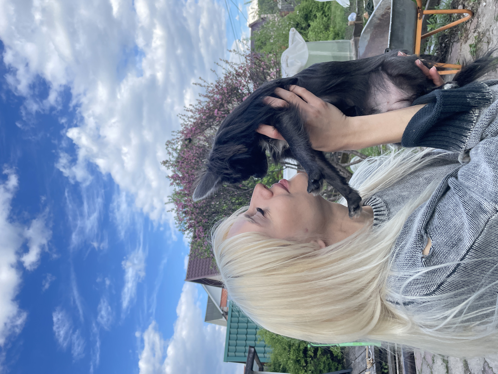 The image shows a woman with blonde hair holding a small, black dog in front of a house. The woman has her face close to the dog. A tree with pink blossoms is visible behind them. The background includes a blue sky with scattered clouds, other houses, and greenery. The location is likely outdoors, possibly in a yard or garden near a residential area in the city of Манский, Russia. The woman is wearing a gray sweater.