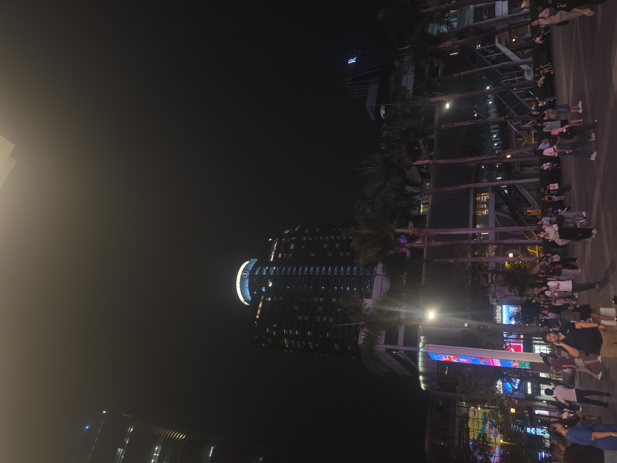 The image captures a vibrant nighttime street scene in Bangkok, Thailand, rotated 90 degrees clockwise. When viewed upright, a wide urban plaza or street is filled with numerous people at the bottom and right side of the frame. Many individuals are seen walking, standing, and conversing, suggesting a bustling evening atmosphere.
The setting is dominated by modern architecture and artificial lighting. A row of tall palm trees lines the main thoroughfare, separating the pedestrian area from what appear to be commercial buildings. Several multi-story buildings rise into the dark night sky. Most notably, a large, dark skyscraper is prominent in the middle-left, featuring numerous illuminated windows and a distinctive, brightly lit "I" logo near its top, likely indicating an InterContinental hotel. Another building further to the right (when upright) displays a prominent blue "R" logo.
The scene is well-lit by various sources, including streetlights, storefront illumination, and large digital billboards. One digital display clearly shows the word "Emirates" in white text, and "MINIWOW" below it. Other colorful, bright signs and screens are visible but their text is largely unreadable. The overall impression is that of a lively commercial or entertainment district after dark, with people enjoying an evening out. The weather appears clear, with no visible precipitation, though the bright lights create some atmospheric haze.