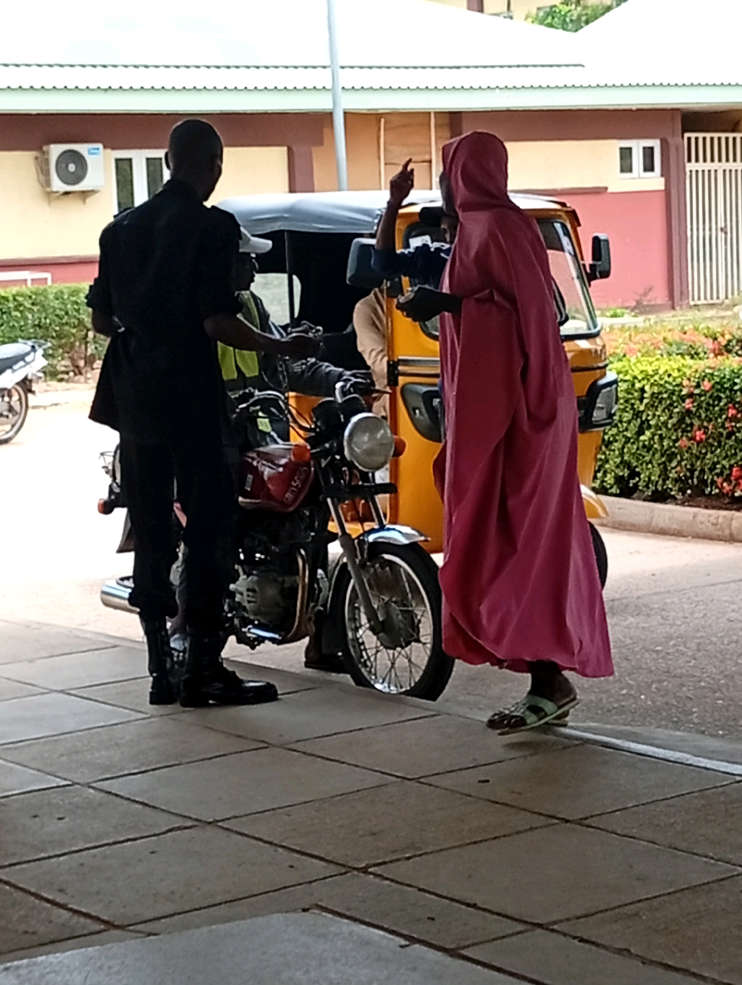 In Gombe, Nigeria, a scene unfolds by a building with a tiled facade and a white roof. In the foreground, two individuals are engaged in an interaction near a yellow and grey auto rickshaw. One individual, wearing black pants and a black shirt, stands with their back to the camera, gesturing with their left hand. Next to them, another person is seen from behind, wearing a pink hooded garment that drapes over their body and feet, which are clad in light green sandals. This individual's right hand is raised, pointing upwards.
Behind them, the auto rickshaw is parked, its front wheel visible. A motorcycle, partially obscured, is parked to the left. The ground is paved with square tiles. In the background, a portion of a building with windows and a security fence is visible, alongside lush green bushes with small red flowers. An air conditioning unit is mounted on the building's exterior. The scene suggests a street-level encounter or transaction in a public area.