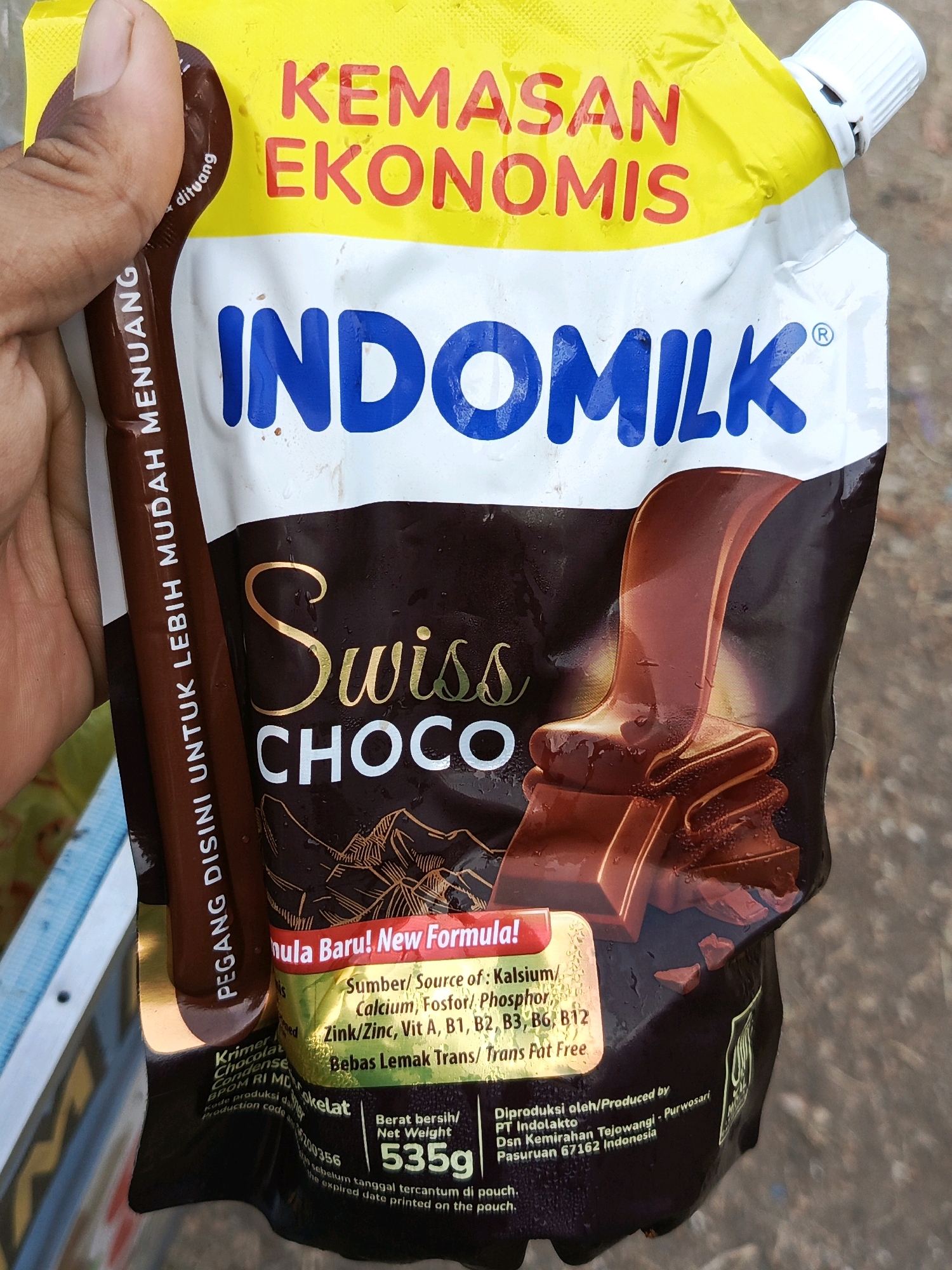 A person's hand is holding a pouch of Indomilk Swiss Choco flavored milk. The pouch is yellow and white with a dark brown bottom that features an illustration of melting chocolate. Text on the pouch includes "KEMASAN EKONOMIS," "INDOMILK," and "Swiss Choco." There is also information about the product's new formula, nutritional content, and production details. The background is blurred and appears to be an outdoor setting, possibly in the city of Megang Sakti II, Indonesia, as indicated by a label on the pouch.