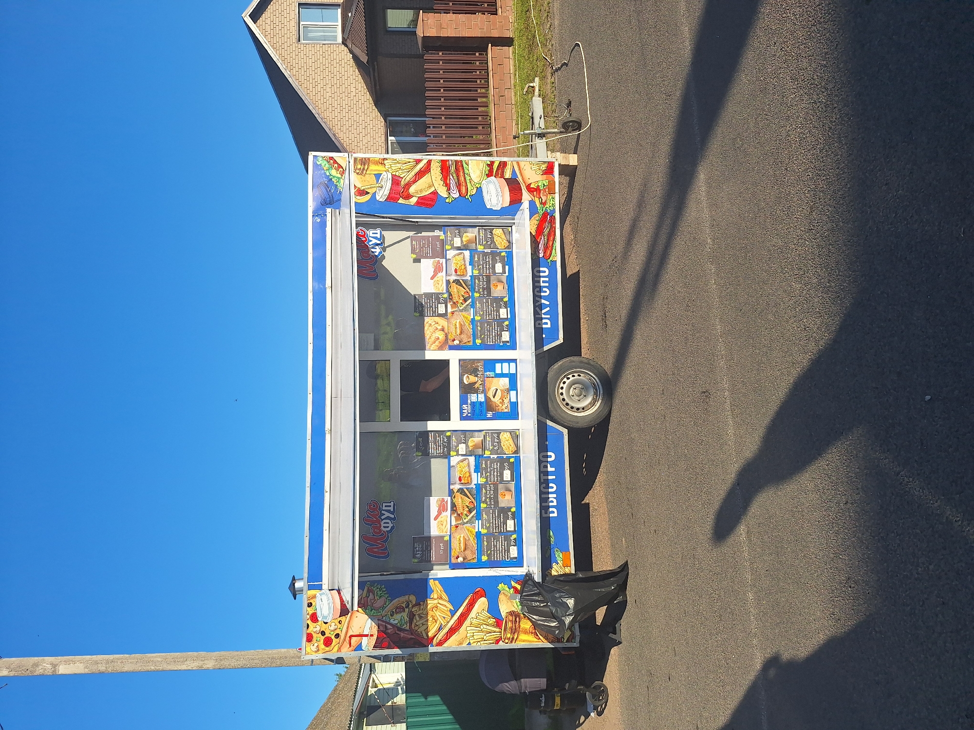 An outdoor, eye-level, full shot shows a blue and white food truck parked on an asphalt road during the day. The food truck is adorned with colorful graphics of food items like pizza, hot dogs, and fries, and features a menu displayed on its side. The words "Make фуд" and "Быстро" are visible on the truck. A person is partially visible inside the truck, likely serving customers. To the left of the truck, a black trash bag lies on the ground next to a small cart with wheels. In the background, a brick house with a brown fence is visible, and a wooden utility pole stands to the left. The sky is clear and blue. The location is Цімкавіцкі сельскі Савет, Belarus.