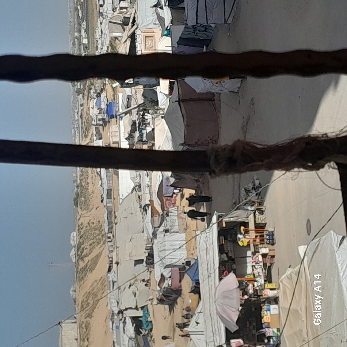 A view of a densely populated camp, possibly for refugees or displaced persons. The camp consists of numerous makeshift tents and shelters, some with laundry lines strung between them. A couple of figures are walking on a pathway or road. The image is shot from an elevated position, looking down on the camp. The foreground is out of focus, with some structural elements partially obscuring the view. The setting appears to be arid or desert-like, and the overall impression is one of crowded and temporary living conditions.