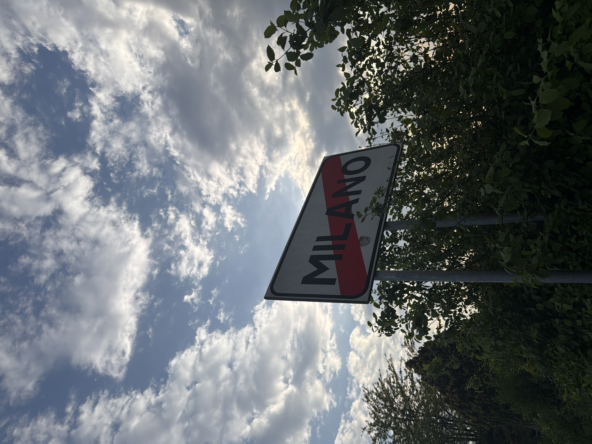 The photograph shows a road sign indicating the end of the municipality of Milan, Italy ("MILANO" written on the sign). The sign is angled upwards towards a sky with scattered clouds. Vegetation, possibly trees and bushes, frames the right side of the image, partially obscuring the sign. The lighting suggests it is daytime.