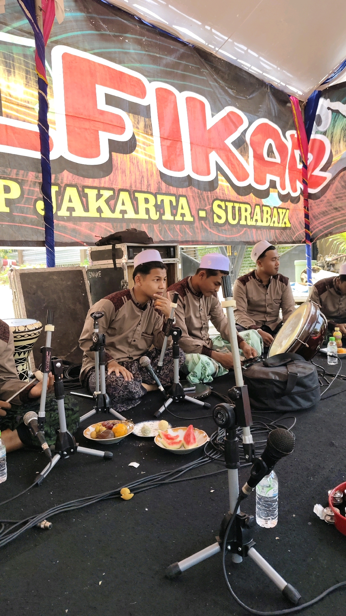 Four young men in light brown traditional clothing and white skullcaps are seated on a black carpeted stage. Two of the men are in the foreground, with microphones and stands positioned in front of them. The man on the left is looking to his right and has his hand near his chin. The man next to him is looking towards the right and appears to be playing a small drum. Behind them, two other men are seated, and one of them has a large drum in front of him. There are plates of fruit, water bottles, and audio equipment scattered on the stage. A large banner with red and white lettering is visible behind the men. The scene appears to be an outdoor event, possibly a performance or religious gathering, given the clothing and the presence of musical instruments and microphones. The banner indicates the event may be related to "FIKAZ" and locations such as "JAKARTA - SURABAYA." There is no specific visual information to confirm the exact location within Banyubunih, Indonesia, beyond it being an outdoor stage event.
