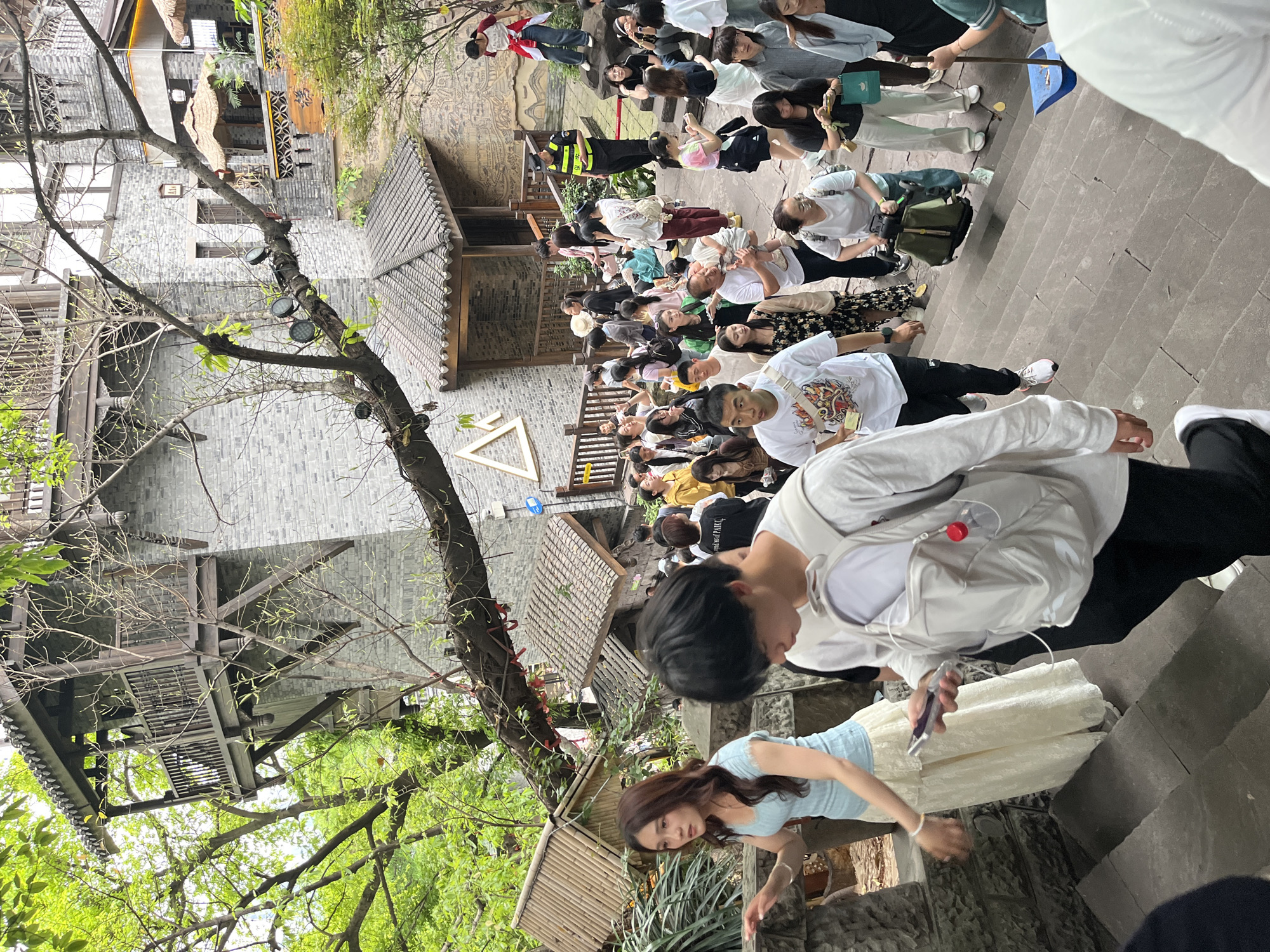 This vertical image depicts a crowded outdoor scene in what appears to be a historical or cultural district in Yuzhong District, China. The setting features traditional Chinese architecture, characterized by grey brick walls, dark tiled roofs, and multi-story wooden structures with balconies. Lush green foliage from trees and plants is visible, framing parts of the buildings.
Numerous people, mostly young adults, are gathered, creating a dense crowd that spans across stone steps and a narrow walkway. Their body language suggests they are either queuing, waiting, or slowly moving through the area, with many looking towards the same direction or observing their surroundings. They are dressed in casual modern clothing.
Notable individuals include a security guard in a yellow and black uniform with the Chinese character "保安" (Bǎo'ān, meaning Security) visible on the back. Another person in the foreground holds a blue dustpan. Many individuals are holding mobile phones, possibly taking photos or interacting with their devices. In the foreground, a person's backpack has a red, circular object attached, possibly a small fan or speaker. On a black t-shirt, partial text "LIVE ON PARADISE" is visible.
The lighting is bright and even, indicating daytime, likely late morning or afternoon on an overcast day, as there are no harsh shadows. The overall atmosphere appears lively and bustling, characteristic of a popular public or tourist area. A white triangular sign is visible on the ground level, potentially a marker or logo.