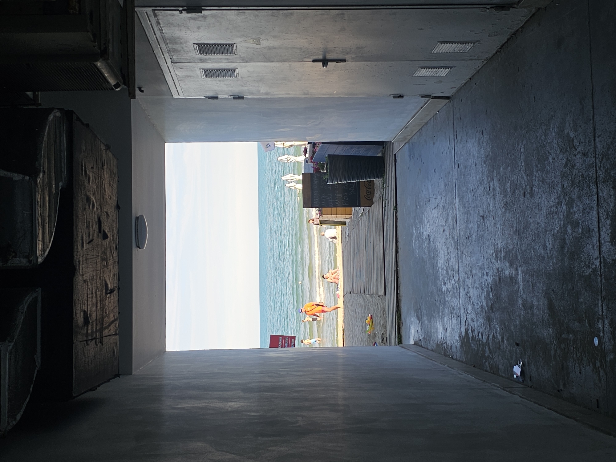 The image depicts a view through a concrete and wooden walkway towards a beach and the ocean. The walkway has rough concrete flooring with some wet patches and a few pieces of litter. On the right, a large grey metal electrical cabinet with two doors and ventilation slats is visible. On the left, a concrete wall forms the side of the walkway.
The walkway opens to a beach with a wooden boardwalk and sand. People are present on the beach, some in the shallow water. One person in an orange shirt and shorts is standing on the boardwalk near the water. Further back, beach umbrellas and some seating areas are visible, suggesting a recreational area. The sea is calm and blue under a clear sky. The overall environment appears to be a transition zone between an urban or service area and a public beach.