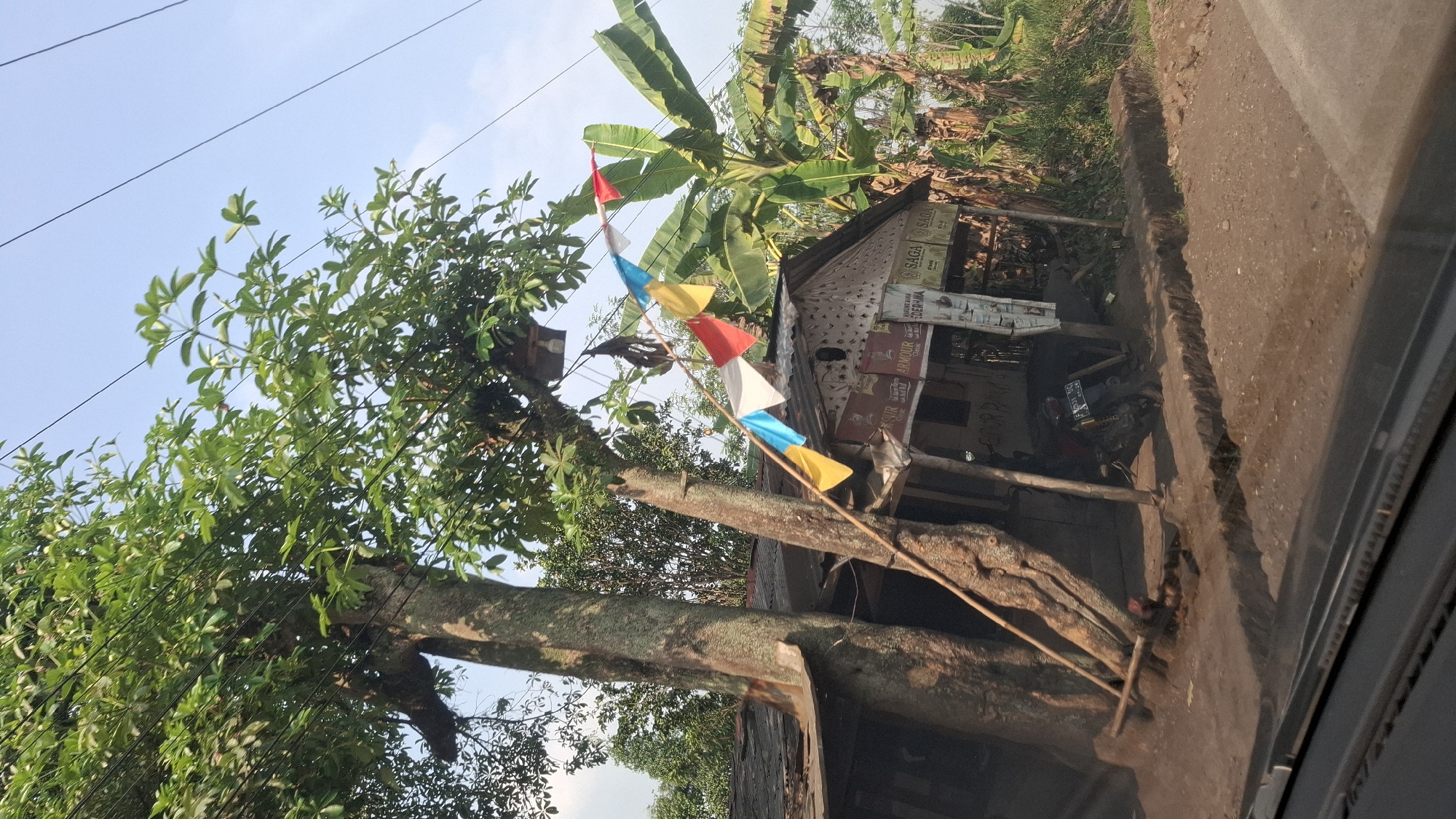 This is a medium shot, eye-level, outdoor, daytime, full-color, documentary-style photograph of a small roadside warung (shop) in Sirnagalih, Indonesia. The warung has a corrugated metal roof and is supported by wooden posts. A large tree with green leaves dominates the left side of the frame, its trunk leaning against the warung. Several small, colorful flags are strung across the front of the warung. Behind the warung, a large banana tree is visible, as well as other lush green foliage. A motorcycle is parked to the right of the warung entrance. The ground in front of the warung is dirt with a small ditch. Power lines run across the top of the frame against a clear blue sky. The image appears to have been taken from a moving vehicle, as the bottom edge of the frame shows the dashboard or interior of a car.