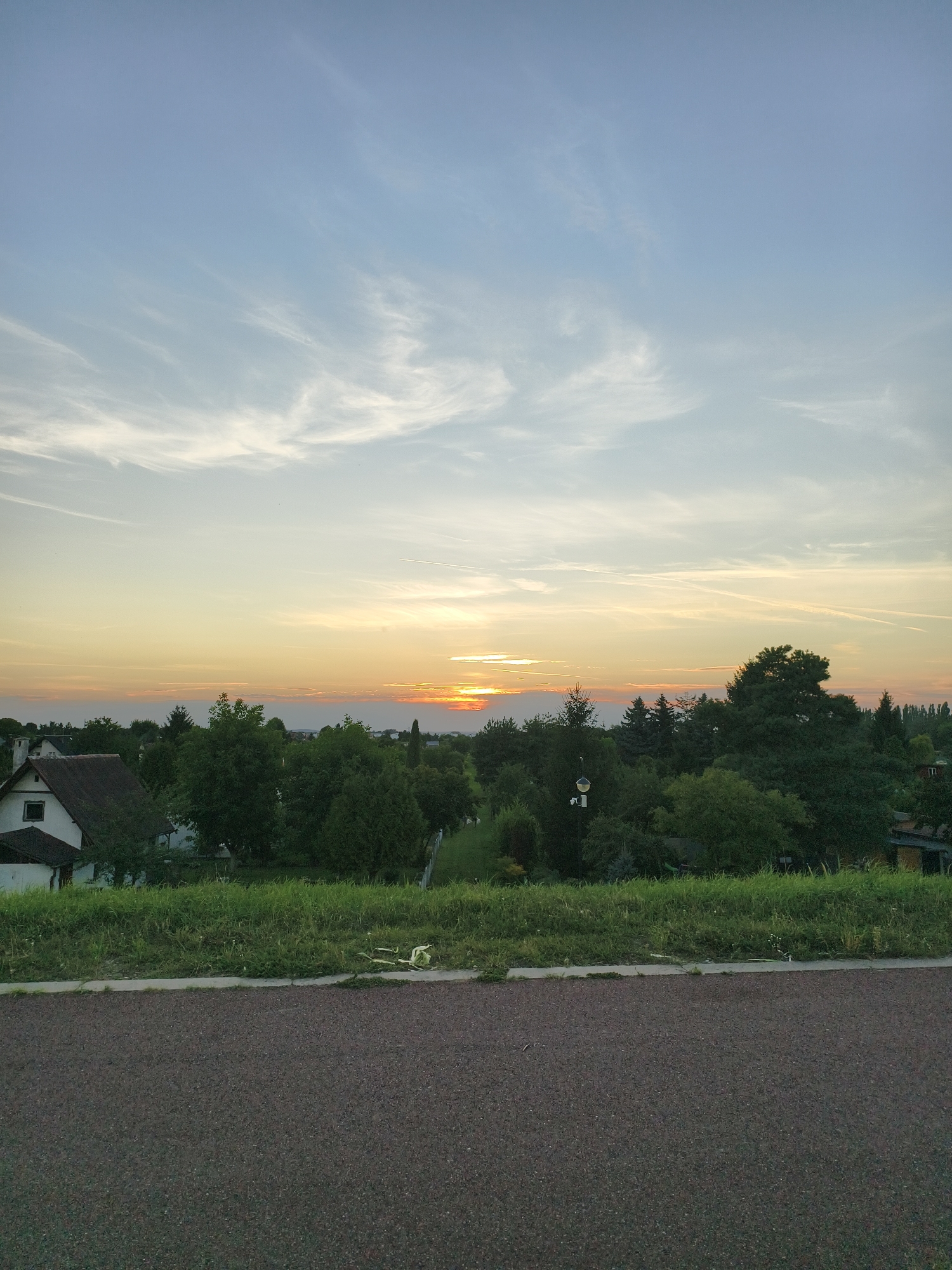 The image depicts a rural landscape at sunset. In the foreground, a dark asphalt surface transitions to a grassy embankment bordered by a concrete curb. Beyond the embankment, a collection of houses and trees are visible. The houses appear to be of modest size with pitched roofs. A dense growth of trees, primarily deciduous and coniferous, forms a significant portion of the mid-ground. The sky above is a blend of pale blue and orange hues, with wispy white clouds stretching across the horizon, indicating the sun has recently set or is about to set. The scene suggests a tranquil evening in a suburban or rural setting. While the image is identified as being in Tczew, Poland, there are no specific landmarks or identifiable features that definitively confirm this location. No people or animals are present in the image.