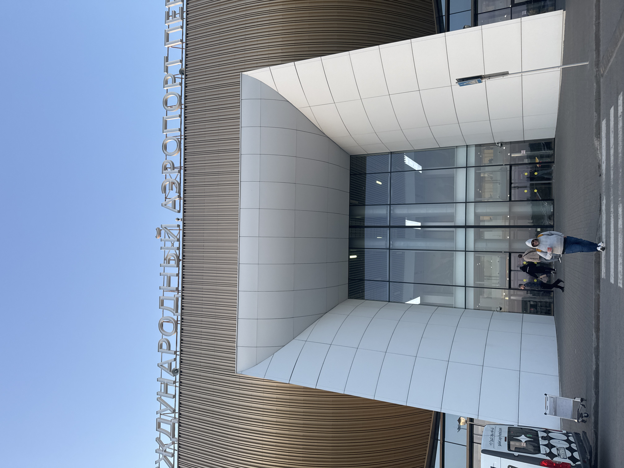 This image shows the entrance of what appears to be the Platov International Airport in Kitsjanovo, Russia, based on the Cyrillic text visible on the building's facade. The architectural style of the entrance includes a modern design with a combination of curved wooden panels and rectangular white panels. A glass facade is centered in the entrance. Several individuals are walking into or out of the airport. A white van with distinctive black and white patterned graphics is parked near the entrance with a luggage cart next to it. The sky is clear and blue.