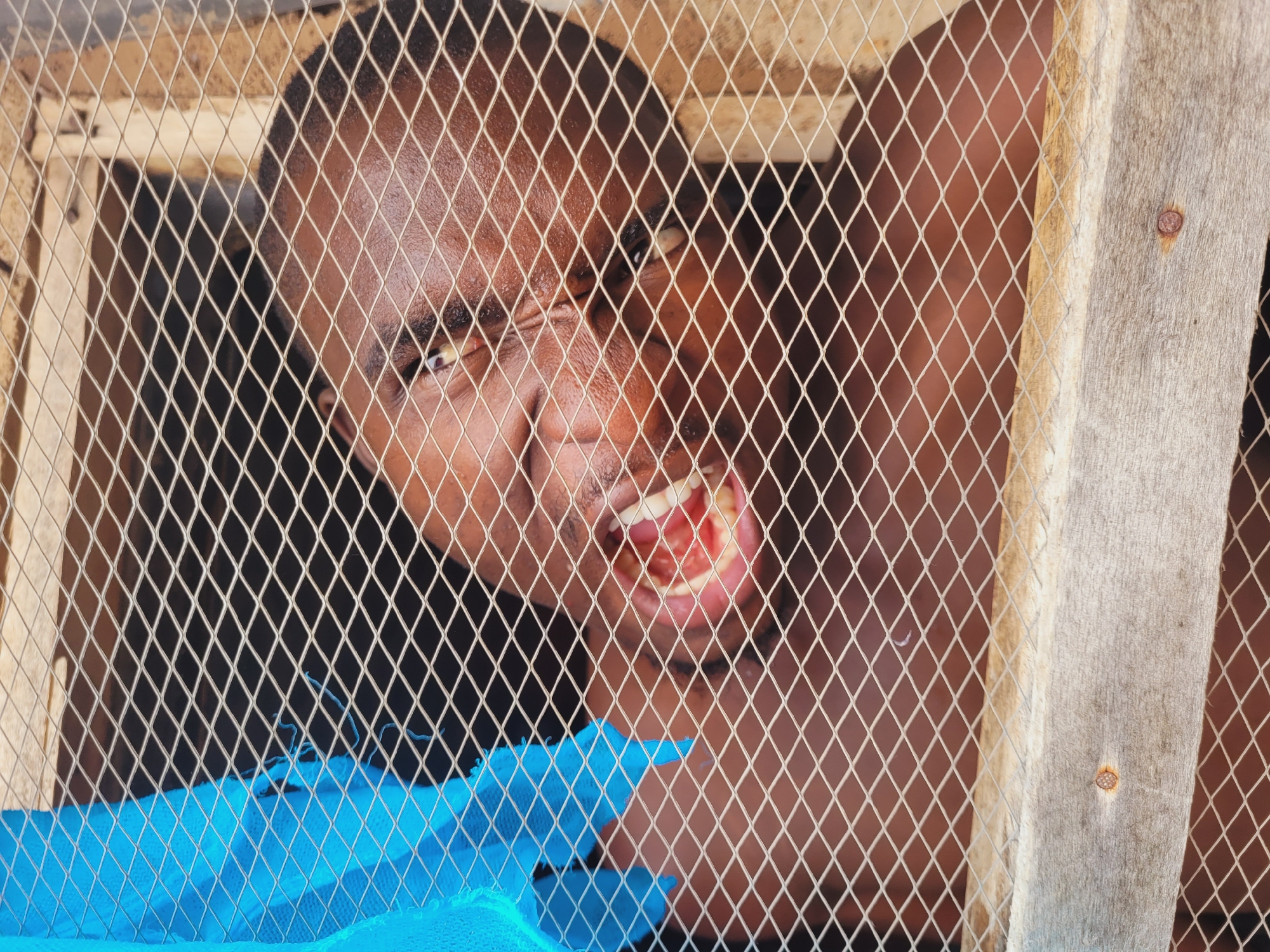 The image shows a close-up, eye-level view of a person's face, obscured by a wire mesh, looking through it. The person's mouth is open, revealing their teeth and gums, and their eyes are visible. A bright blue, textured material, possibly fabric or plastic, is partially visible on the left side of the image. The background is dark, with some wooden elements and more of the wire mesh. The scene suggests a confined space or a barrier between the viewer and the subject. The location in Bukuru, Nigeria, implies a specific setting within that city, but the image itself does not contain explicit geographical markers.