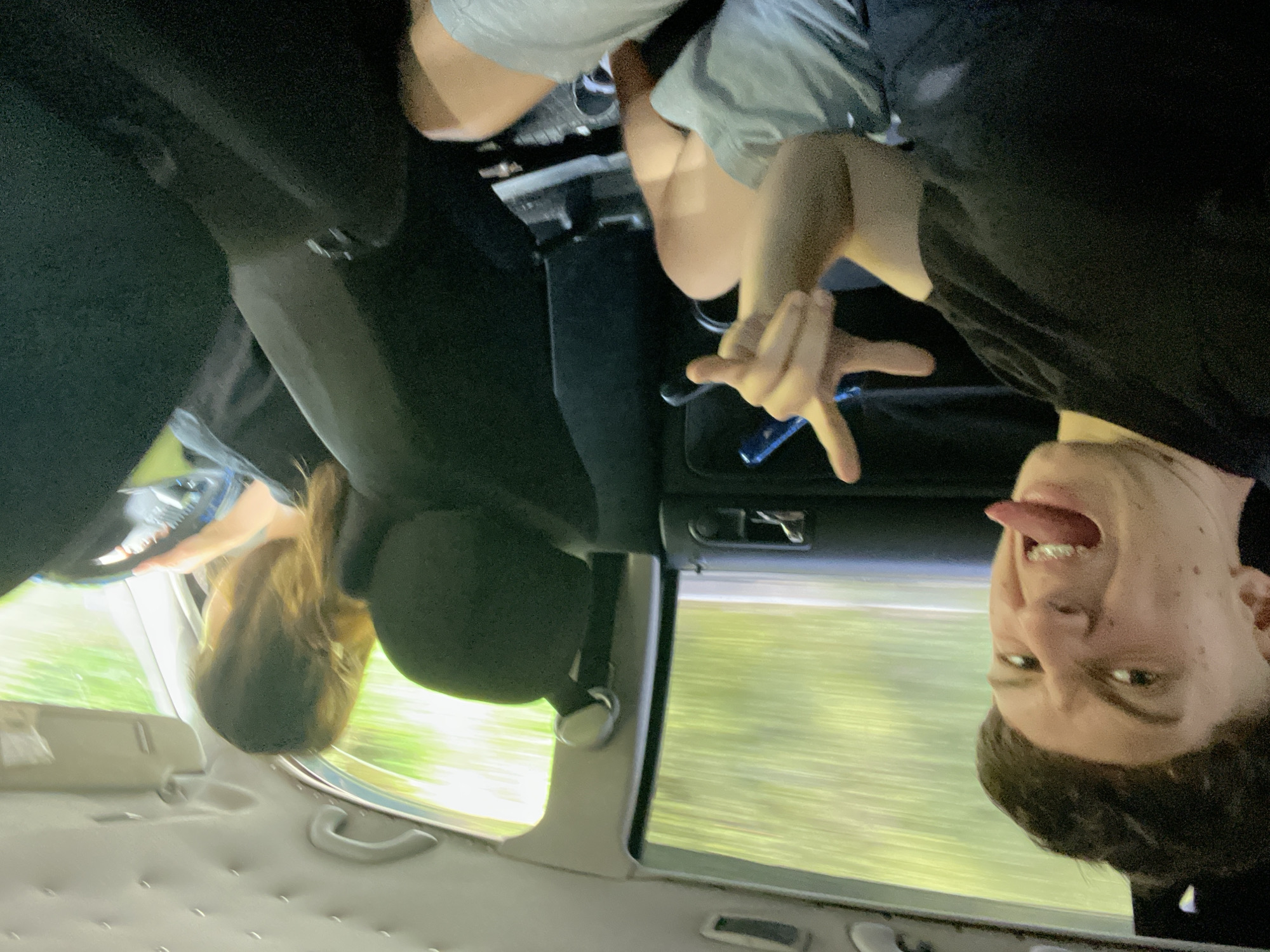 This is a candid, medium-shot, eye-level photograph taken from inside a moving vehicle. The perspective is looking towards the front of the car. In the foreground, a young Caucasian male with short, styled dark hair is making a face at the camera, sticking out his tongue which has braces. He is wearing a black t-shirt and grey shorts, and is positioned on the left side of the frame. His right hand is raised, with two fingers extended in a peace sign, and he is holding a blue object. Behind him, to the right and slightly blurred, is a young Caucasian female with long, light brown hair, who appears to be driving. She is wearing a black top and is holding the steering wheel. The interior of the car is visible, including dark upholstery and the car's ceiling. The windows show a blurred background of green foliage, suggesting the vehicle is traveling on a road surrounded by trees. The lighting is natural and bright, indicative of daytime. There is no discernible information to place this image specifically within the city of Vlahovo, Bulgaria, or any other specific urban location.