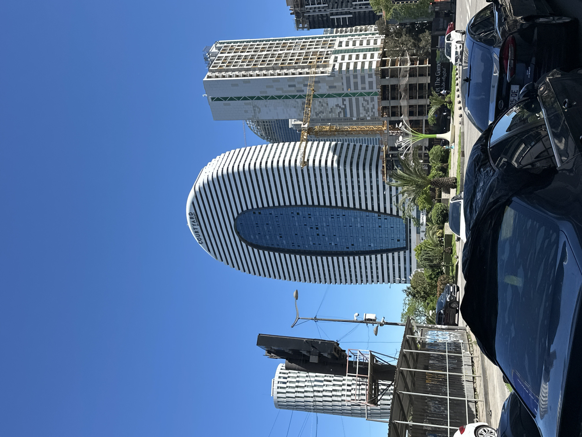 The image captures a scene in Batumi, Georgia, featuring a modern, oval-shaped high-rise building with white and dark blue horizontal stripes. The building is likely the Courtyard by Marriott hotel due to the logo on the side. In the background are other high-rise buildings, one still under construction with a visible crane. Street-level elements include palm trees, decorative sculptural features, and parked cars. A partially obscured structure, perhaps a bus shelter or construction barrier, is present in the foreground. The sky is clear and blue, suggesting a sunny day.