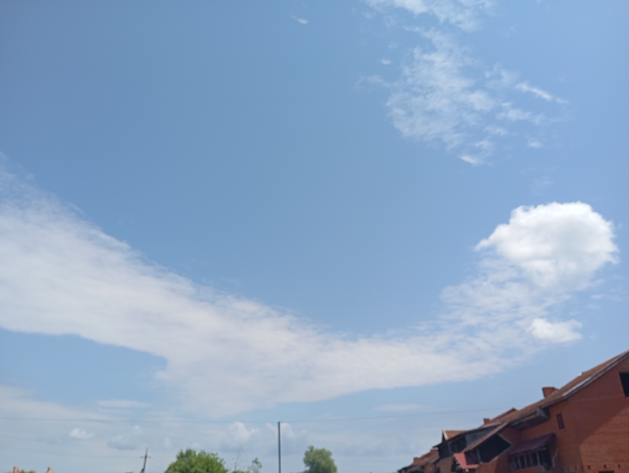 The image shows a clear blue sky with scattered white clouds. In the lower right portion of the image, there are buildings with red brick walls and tiled roofs, suggesting an urban or suburban environment. The architecture appears to be European, possibly in Saratovskaya, Russia, given the hint. The scene is static, with no visible people or action. The overall impression is a bright, sunny day.