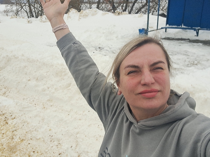A selfie photograph depicts a Caucasian woman in Sasovo, Russia, set against a snowy outdoor backdrop. The woman, appearing in her 30s or 40s, is captured from the chest up, wearing a grey hooded sweatshirt. Her blonde hair frames her face, which displays partially squinted eyes and slightly pursed lips. Faint reddish marks are visible on her cheeks. Her right arm is raised vertically with her hand open, and a light pink segmented wristband is secured around her right wrist. The ground behind her is covered in dirty, partially melted snow, exhibiting brown patches and faint impressions. In the mid-ground and background, bare deciduous trees and mounds of snow are present. To the right of the frame, a segment of a blue structure, possibly metal, resembling a small building or enclosure, is visible, along with a blue bench.