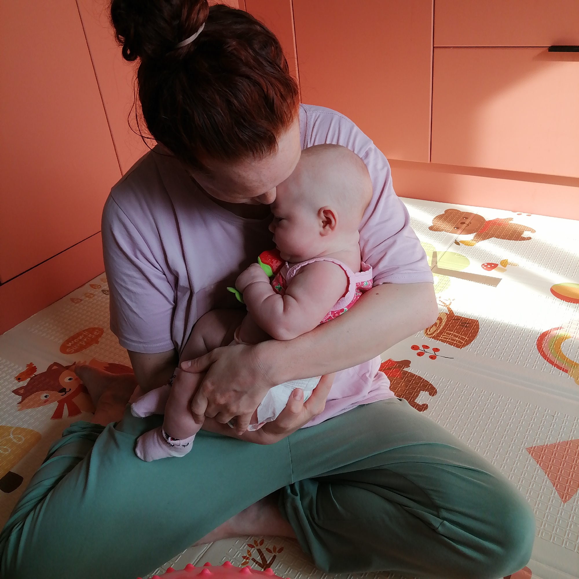 An overhead, medium shot captures a woman holding a baby on her lap. The woman, with auburn hair pulled into a messy bun, wears a light pink t-shirt and green pants. Her head is bowed, resting against the baby's head, and she cradles the infant in her arms. The baby, dressed in a pink onesie and a diaper, is held close, with its face turned away from the camera. The baby's right hand holds a red and green teething toy. They are seated on a light-colored play mat adorned with cartoon animals and shapes, placed on a wooden floor. The background features coral-colored cabinetry with a black handle, suggesting an indoor domestic setting. The scene appears to be in a residential environment, possibly a child's room or play area. The presence of a play mat and a teething toy indicate the child's young age. The overall lighting is warm and diffused, with a bright light source casting shadows on the play mat. The specific location of Trubacheyevka, Russia, is not discernible from the visual elements of the image itself.