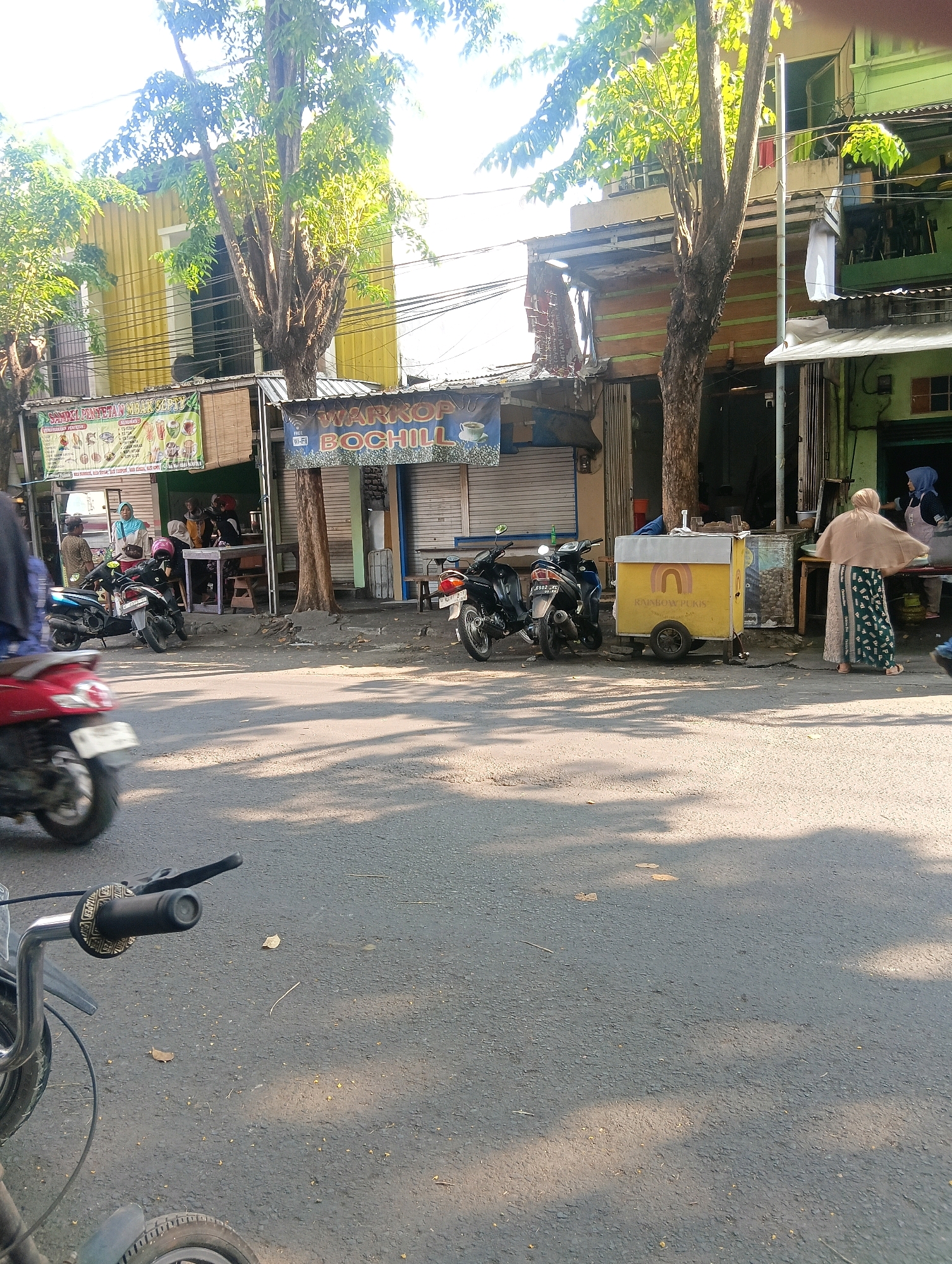 The image captures a street scene in Gapurosukolilo, Indonesia. In the foreground, a dark asphalt road is visible, with shadows of trees cast upon it, indicating it is daytime. A bicycle handlebar and front wheel are prominently featured in the lower left corner, suggesting the perspective of someone on a bicycle.
Further down the street, several parked motorcycles are lined up on the side of the road. To the right of the motorcycles, a yellow food cart with a rainbow logo and the text "RAINBOW PURIS" is positioned. Behind the cart and motorcycles, a row of shops and buildings lines the street. One shop has a banner displaying food items and drinks with text in Indonesian. Another shop has a sign that reads "WARKOP BOCHILL" with a Wi-Fi symbol.
Several individuals are present. In front of the "WARKOP BOCHILL" shop, a woman wearing a beige hijab and a patterned skirt is standing, seemingly interacting with someone inside the shop. Another woman in a blue headscarf and apron is also visible behind a counter. Inside the shops, more people can be seen seated at tables. A red scooter is partially visible on the far left, moving away from the camera. The buildings are a mix of styles, with some painted in bright colors like yellow and green, and others appearing older with weathered facades. Trees with lush green foliage are scattered throughout the scene, contributing to the urban, yet somewhat tropical, atmosphere.