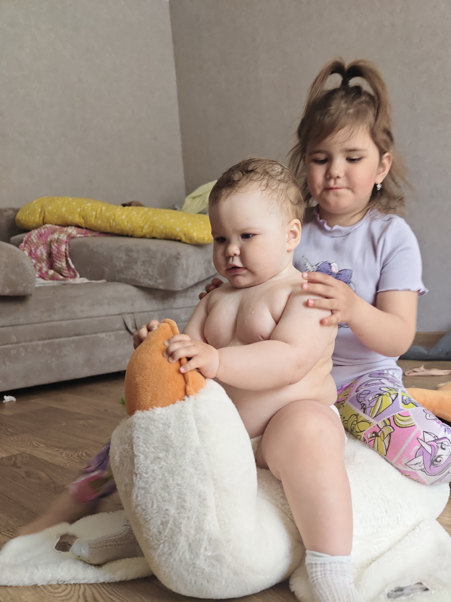 The image depicts two young girls in an indoor setting, likely a home. The younger child, who appears to be a baby, is seated on a plush toy that resembles a horse or a similar animal. She is wearing white socks, but her upper body is bare. The older child, with her hair tied up, is kneeling behind her, placing her hands on the baby's shoulders. She is wearing a light purple t-shirt and patterned leggings.
A sofa is visible in the background, draped with a yellow cushion and a pink patterned blanket. The setting appears to be a room in a residential building. The location information suggests this might be in the city of Chaikovsky, Russia.