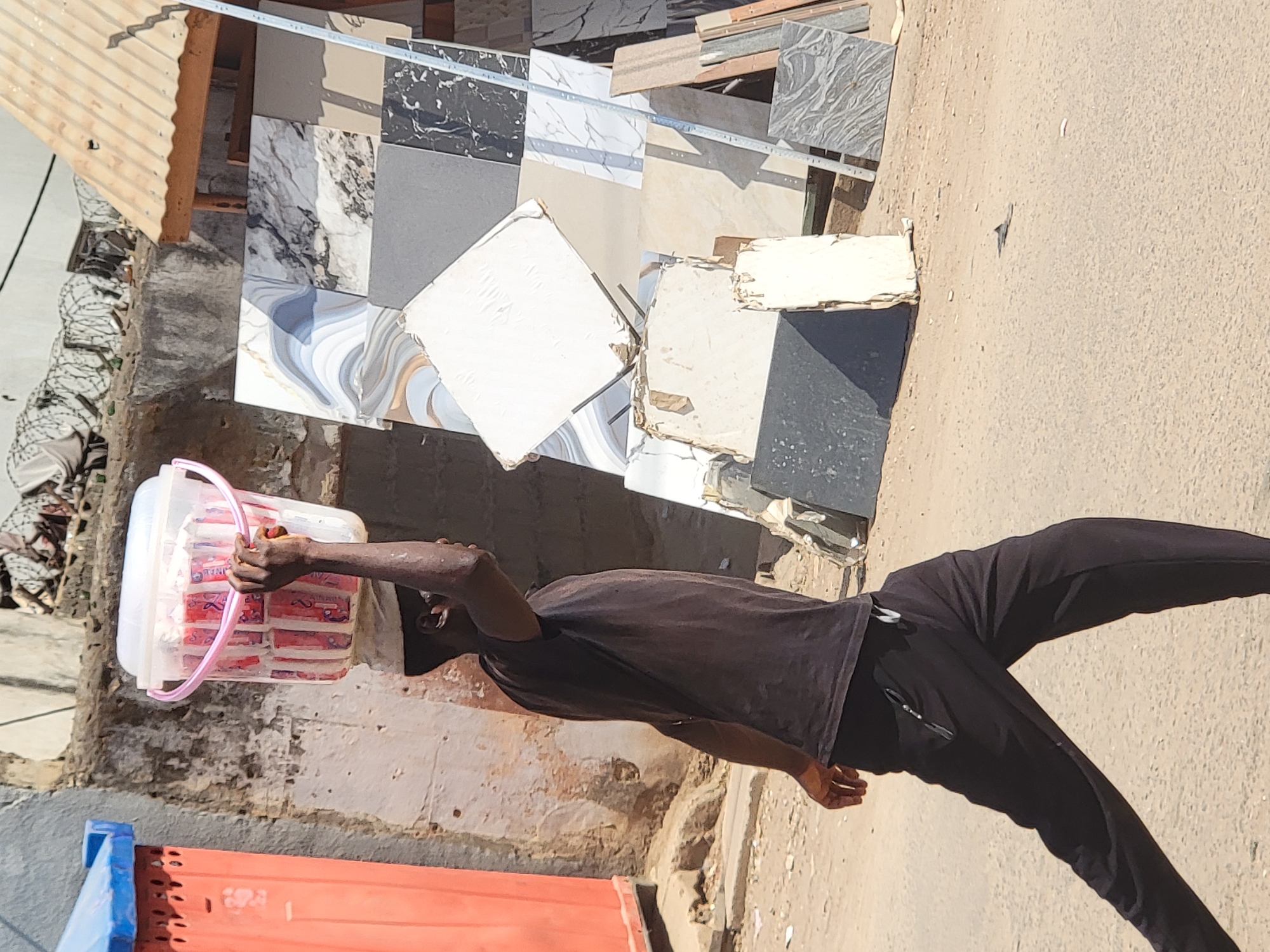 A street scene in Maiburiji, Nigeria, shows a man carrying a white plastic bucket filled with small, rectangular packets overhead, walking on a paved street next to a building displaying numerous tile samples. The man is wearing a dark t-shirt and dark athletic pants. The building is constructed of brick, and is adjacent to a corrugated metal awning. A red and blue bin or container is partially visible on the left edge of the image.