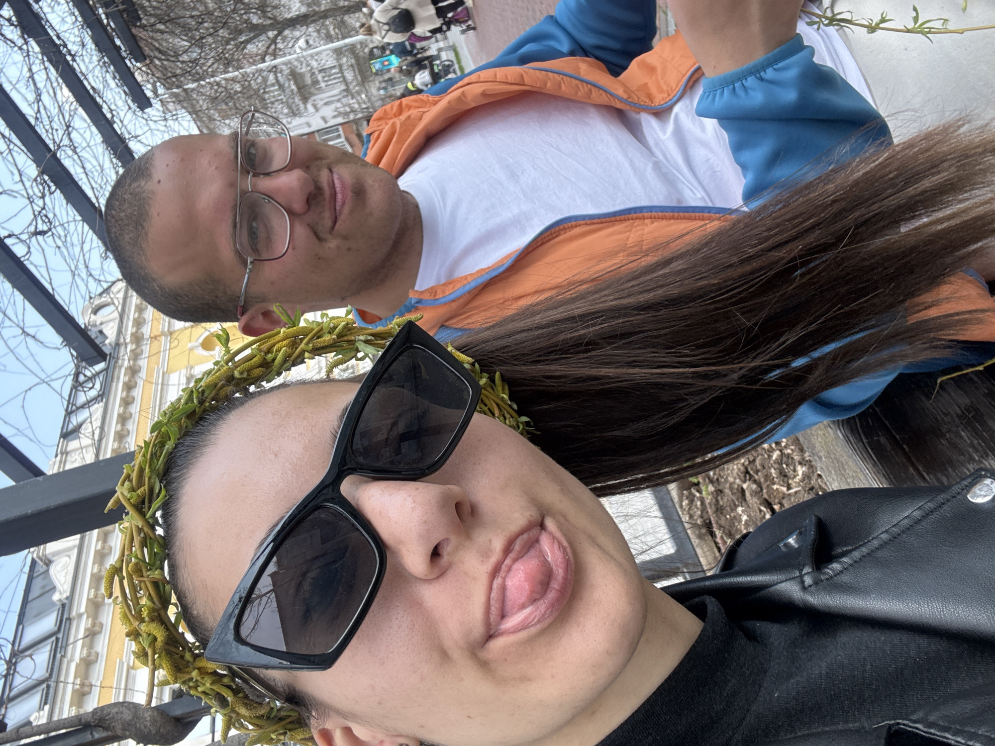 The image shows a selfie of two individuals. A woman with sunglasses and a floral head wreath is sticking out her tongue. She is wearing a black leather jacket. Behind her is a man wearing glasses and a white t-shirt under an orange and blue jacket. In the background, buildings and other people are visible, suggesting an urban setting. The image was taken in the city of Varna, Bulgaria.
